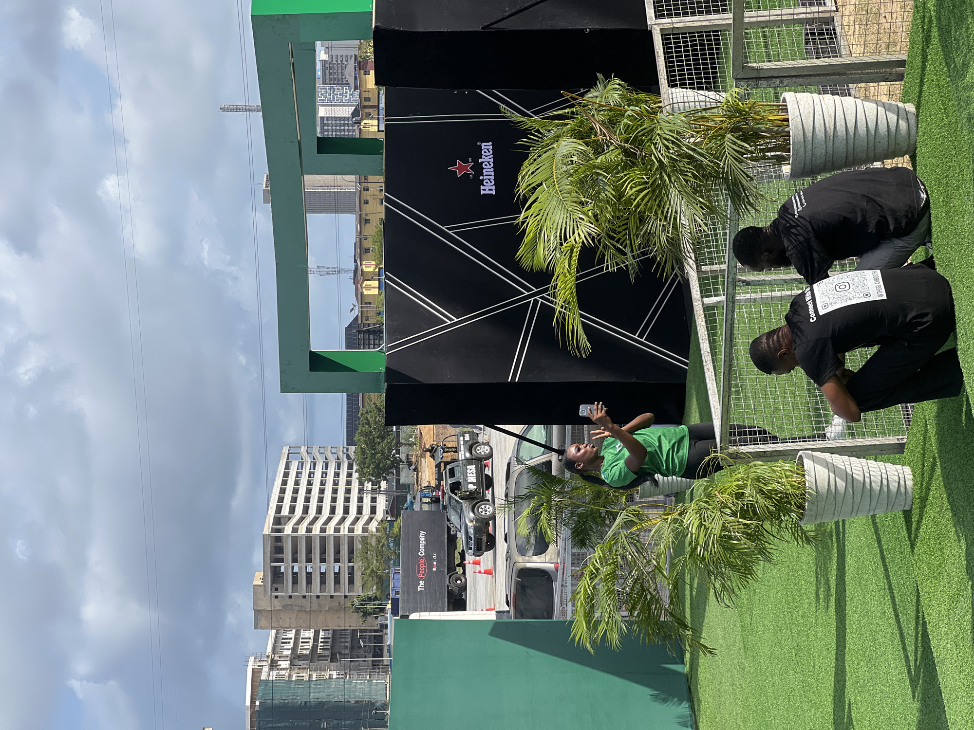 The image shows an outdoor event setup in Lagos, Nigeria, identifiable by the distinctive architecture in the background. A black panel with a white Heineken logo dominates the center of the frame. A person in a green shirt stands near the panel, holding and looking at a smartphone. Two other individuals wearing black shirts are kneeling on the ground near a grey metal fence and potted palm trees. One shirt has a QR code and social media handle on the back. The scene is set on artificial green turf. Behind the event setup, a street scene with various vehicles and a building displaying "The People Company" is visible. Several unfinished buildings are visible in the background sky.