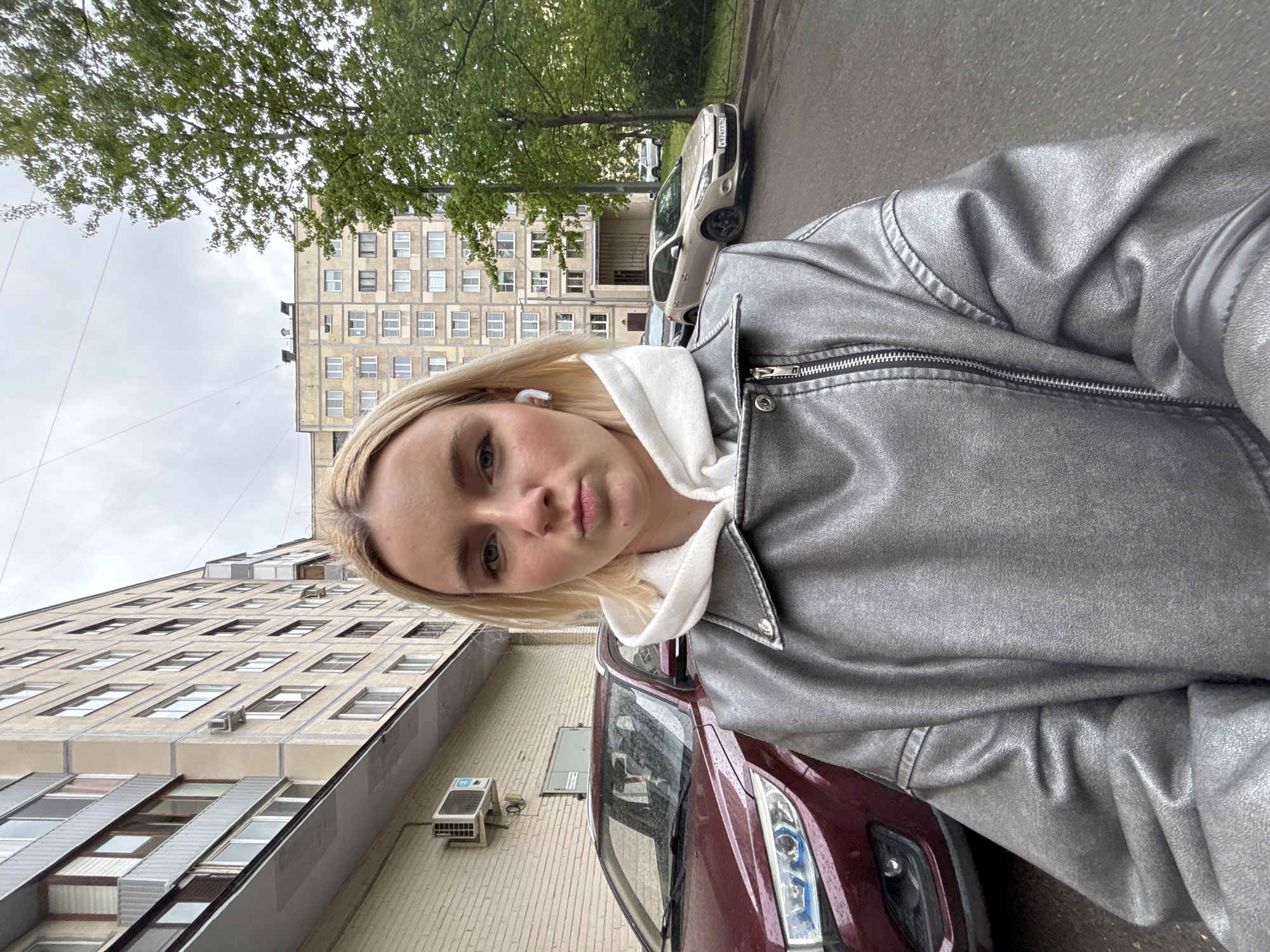 A young woman with blonde hair is taking a selfie outdoors. She is wearing a white hoodie and a silver jacket. In the background are apartment buildings, trees, and parked cars. One of the cars is red, another is silver. The license plate number visible on the silver car is O 977 TT 47. The building facades appear to be in Novoe Devyatkino, Russia.