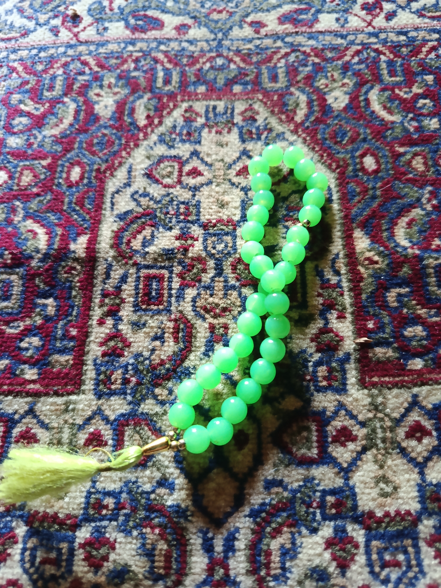 A strand of light green prayer beads with a yellow tassel lies on a prayer mat with a patterned design of red, blue, and cream colors. The beads are round and translucent, strung together on a thin cord. The prayer mat has a repeating geometric pattern characteristic of Islamic art. There are no people or actions depicted in the image. The lighting suggests an indoor setting, possibly during the daytime due to the presence of shadows. The exact location in Karangmojo, Indonesia, cannot be determined from the image itself.