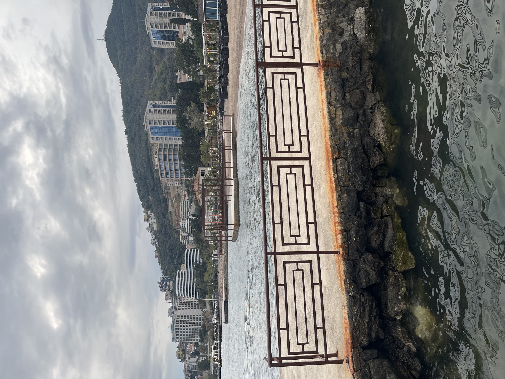 The image shows a waterfront view in Budva, Montenegro. Buildings line a mountain rising from the coast. A patterned metal railing stands along a concrete walkway edging the water. The water surface is rippled, reflecting the cloudy sky. Below the walkway, a rocky edge transitions into the water.