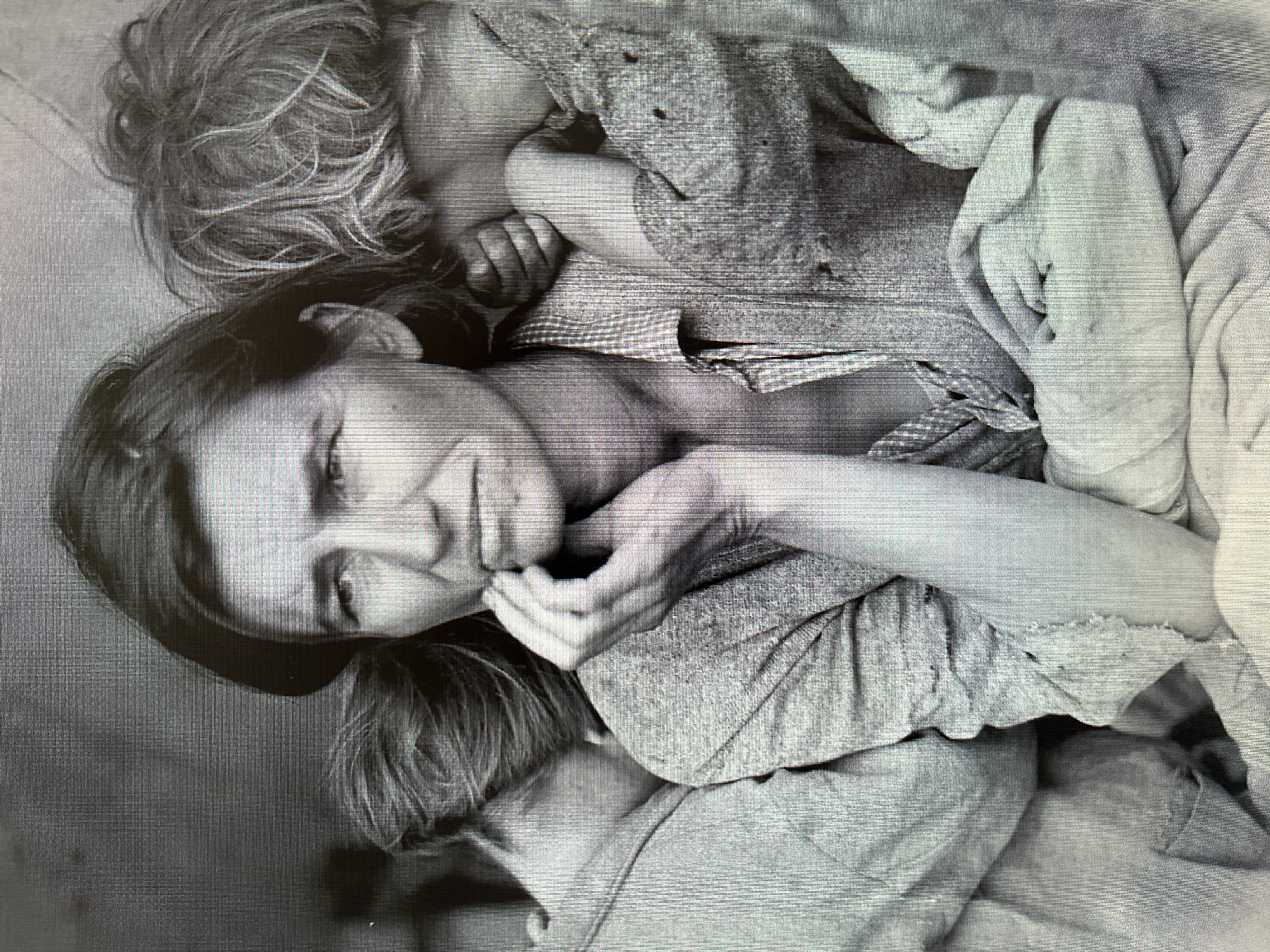 The black and white photograph depicts a woman surrounded by two children. The woman has dark hair, and her hand is resting on her chin. The children, one with blonde hair, are nestled close to her. All three are wearing simple, worn clothing. The setting appears to be a modest interior. There is no indication that this is in Ореховка, Russia, or any city at all.