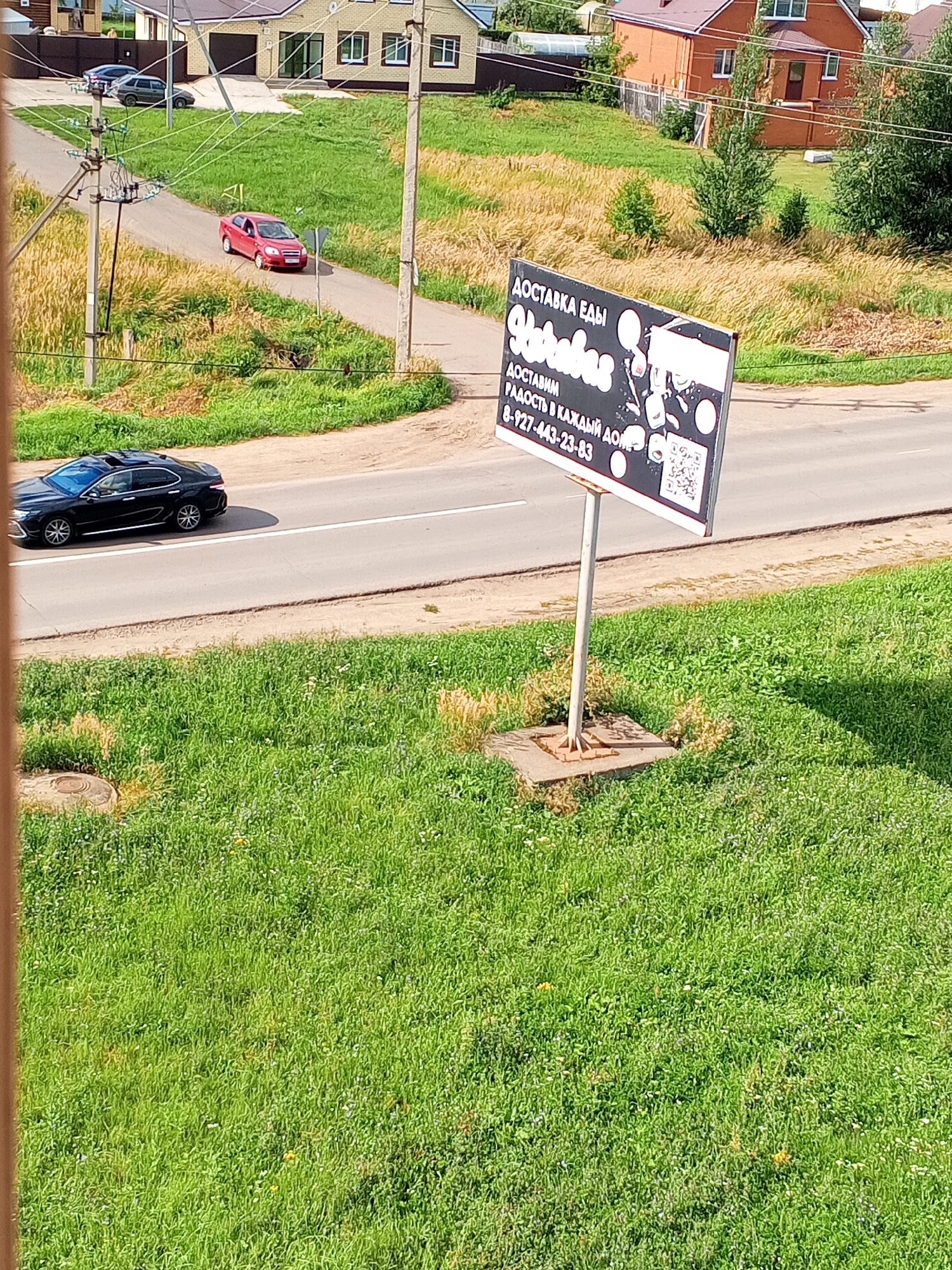 The image shows a street scene in Chistopol, Russia. In the foreground, a black sedan is driving on a paved road. To the left and right of the road is a grassy area. In the background, there is a red car parked on a dirt road. Further in the background, there are several houses and trees. A billboard advertising food delivery is also visible on the right side of the image. The sky is blue and there are some clouds.