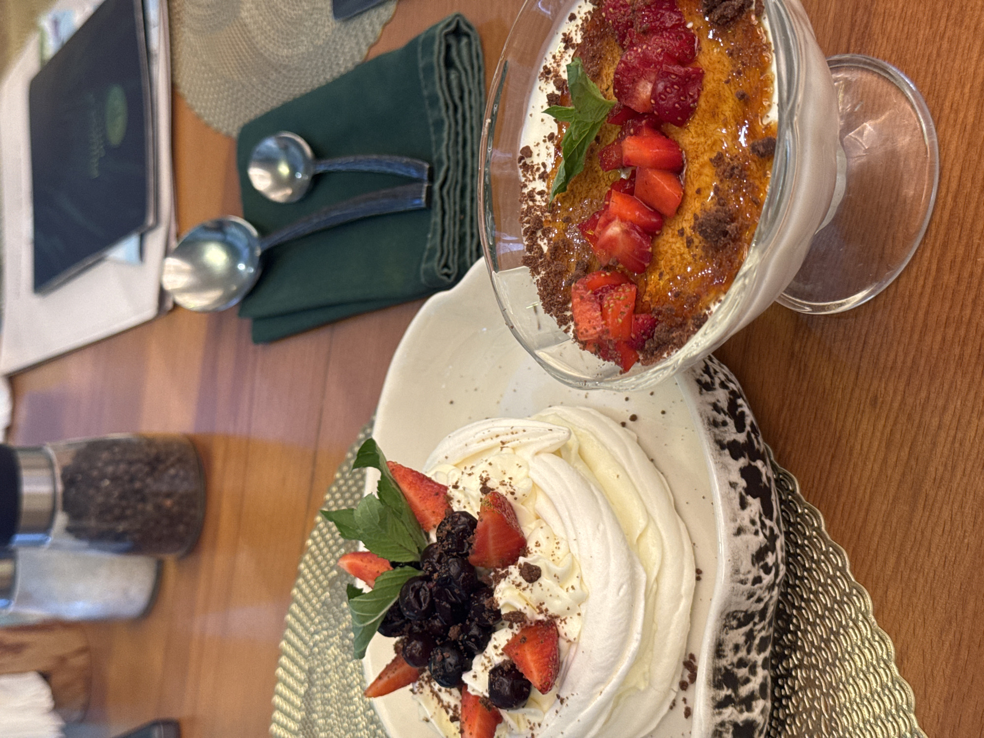 The image shows a close-up, high-angle shot of two desserts on a wooden table. On the left is a Pavlova topped with blueberries and strawberries, dusted with chocolate. It sits on a decorative white plate with a black pattern. To the right is a glass dessert bowl containing a creamy dessert with a brûléed sugar topping, garnished with chopped strawberries and a sprig of mint. In the background, out of focus, are two salt and pepper grinders, a folded green napkin with two spoons, and what appears to be a menu or book. The setting is likely a restaurant or cafe in Sosnoviy Bor, Russia, given the presence of these elements.