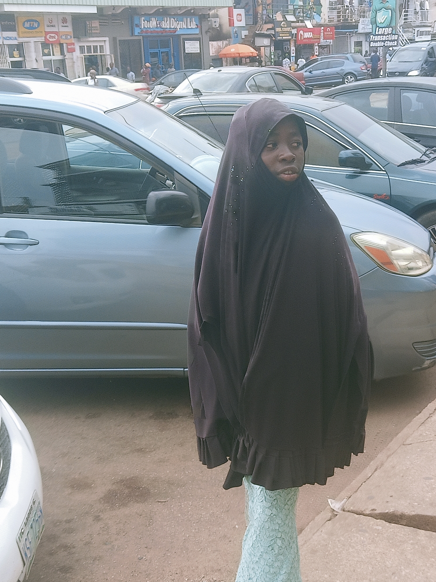 A medium shot captures a person wearing a dark, full-body hijab, speckled with small embellishments, standing on a sidewalk. The individual has their head uncovered, and their lower body is clad in a light blue textured fabric. The scene takes place on a street in Jos, Nigeria, as suggested by the presence of a MTN sign and signage for businesses like "Folowrld Digital Lab" and advertisements reading, "CKEY DU Large Transaction Double-Check". Several cars line the street, with one light blue vehicle prominently positioned in the foreground on the left side, obscuring part of the person from the waist down. The street appears busy, with multiple buildings, signs, and people visible in the background.