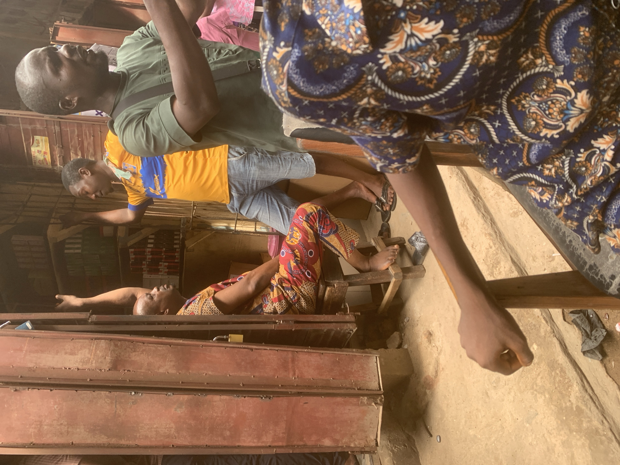 The image, taken at a low angle, depicts a scene in Fegge, Nigeria. In the foreground is a stack of weathered metal coffins. A man lies atop one, his arm outstretched. Another man stands near the coffins, looking at him. Behind them, a man in a yellow shirt is standing and looks at the scene as well. The ground is dirt. A man wearing an ornamental patterned shirt stands on a stool.