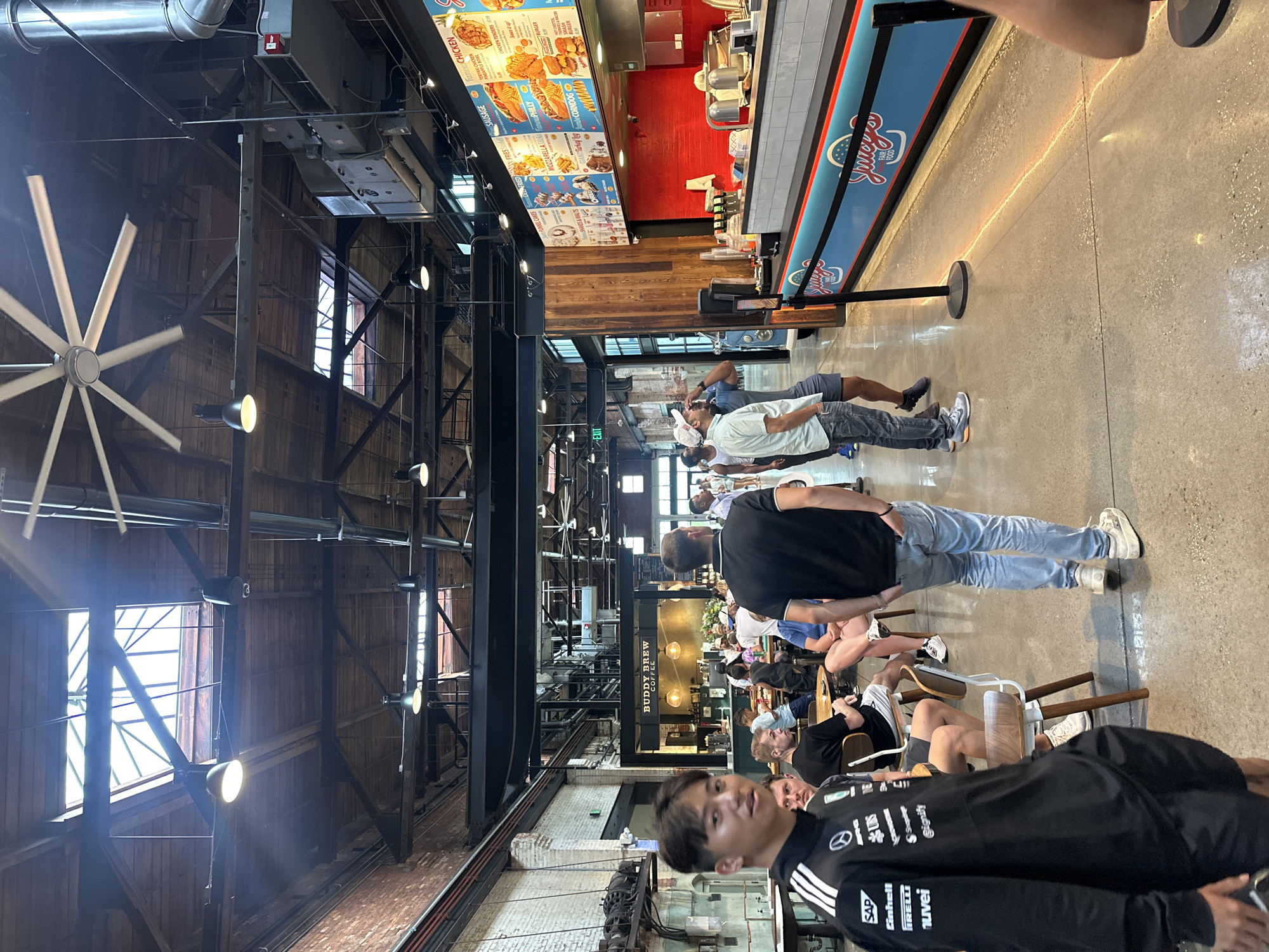 The image captures an interior scene, probably in a spacious industrial-chic dining establishment in Tampa, United States. Patrons stand in line at "Spole's", a casual food establishment which prominently features menu items like chicken, mozzarella fries, and shakes. A group of people are seated at tables in the dining area further inside the building at "Buddy Brew Coffee". The architecture features high ceilings with exposed beams and industrial-style lighting fixtures. A large fan is visible near the ceiling. A man wearing a black sweatshirt with corporate logos faces the camera. People are standing, seated, and waiting in line. The atmosphere appears to be casual and social.