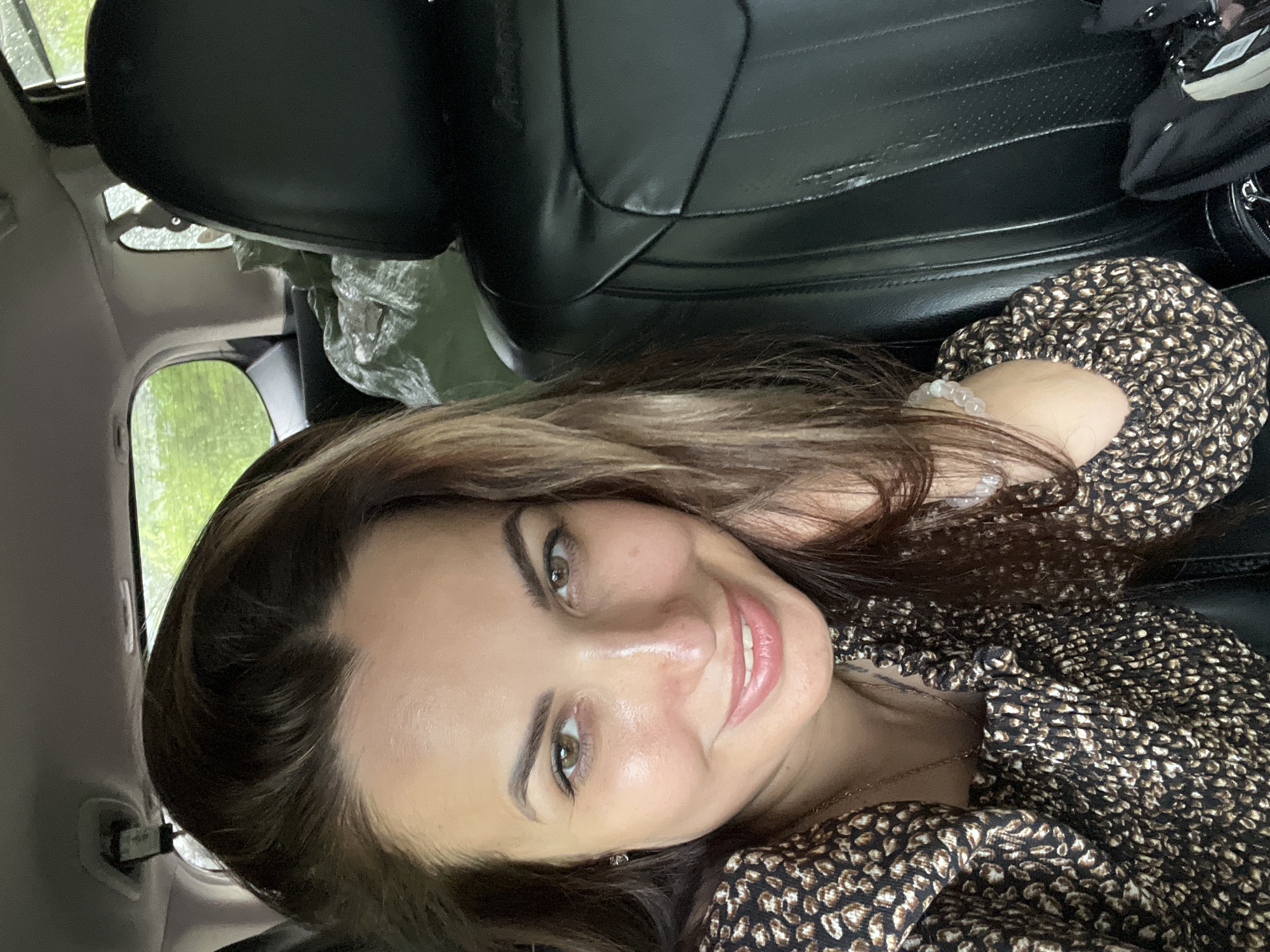 A woman is pictured in the interior of a vehicle. She is smiling directly at the camera, her head tilted slightly to her left. Her hair is dark brown and pulled back, with some strands framing her face. She is wearing a dark top with a tan animal print. To her right, a black leather car seat is visible. In the background, through the car windows, the scene is blurred green foliage.