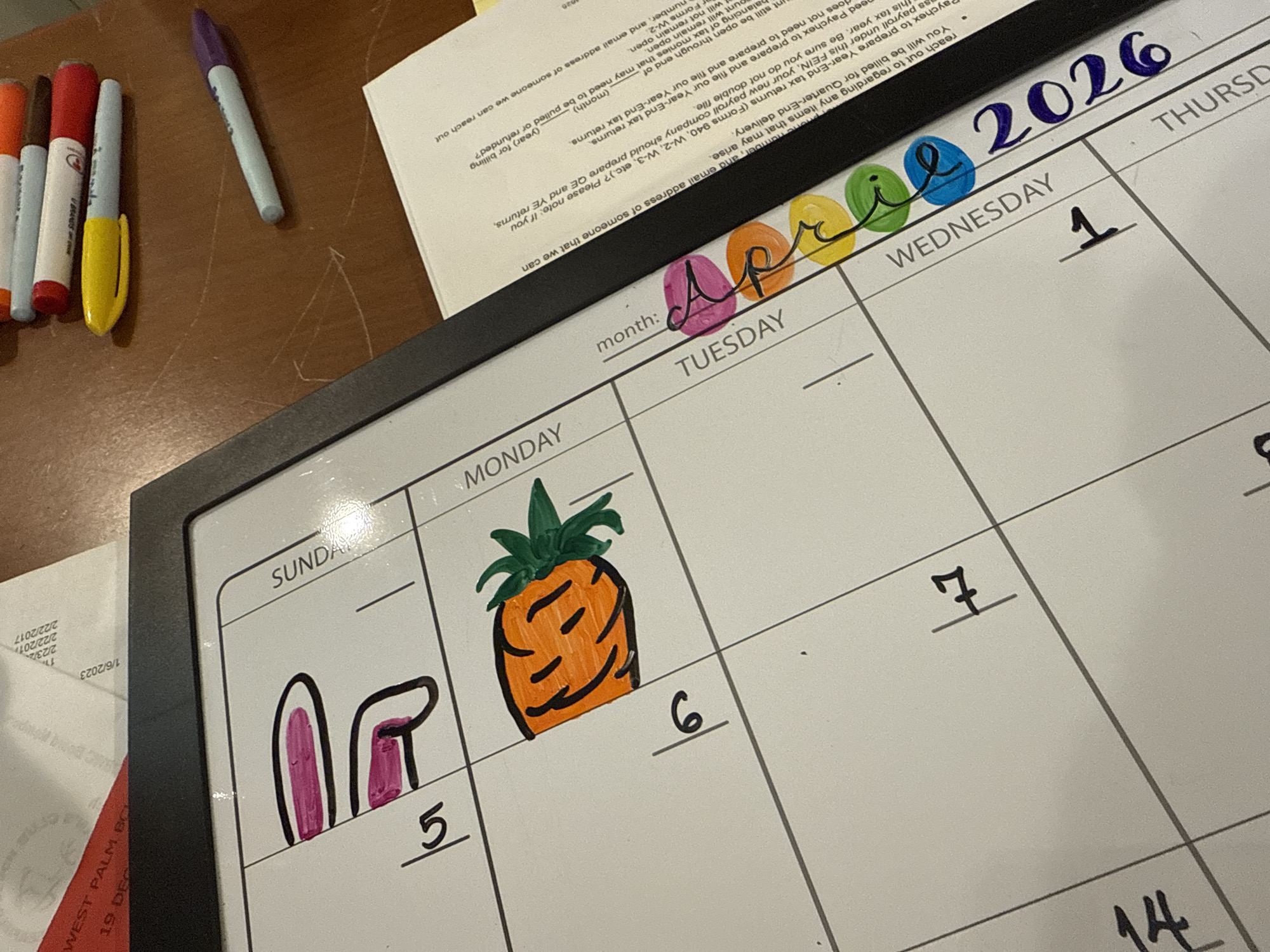 This image shows a calendar open to April 2026, with handwritten text and drawings. The month of April is written in colored markers (pink, orange, yellow, green, blue, and purple). The calendar grid displays the days of the week starting with Sunday, going to Monday, Tuesday, Wednesday, and Thursday. The 5th of April is marked with a drawing of pink bunny ears. The 6th is marked with a drawing of a pineapple. The 1st of April and the 7th and 14th are marked with numbers. The calendar is sitting on a table in Davie, United States of America. Also on the table are various markers and a piece of paper with printed text.