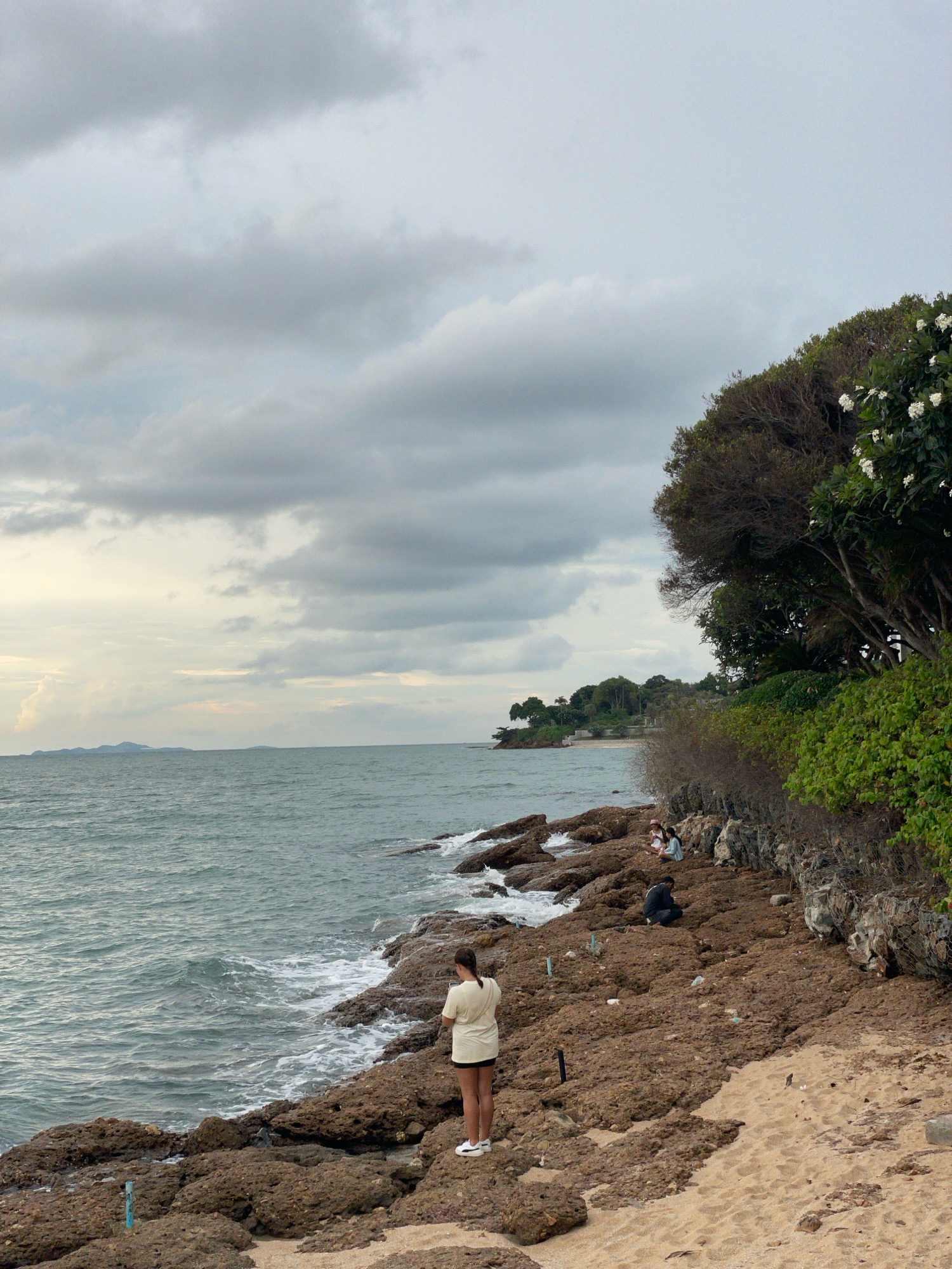 A wide shot captures a rocky coastline in Pattaya City, Thailand, under a cloudy sky. The ocean is on the left, with waves gently crashing against the rocks. On the rocky shore, a woman stands facing the water, wearing a light-colored t-shirt and dark shorts. Further down the shore, several other individuals are seated on the rocks. Lush green vegetation, including trees with white flowers, grows on the elevated embankment to the right, obscuring a portion of the horizon. The ground transitions from rocks to a sandy beach in the foreground.