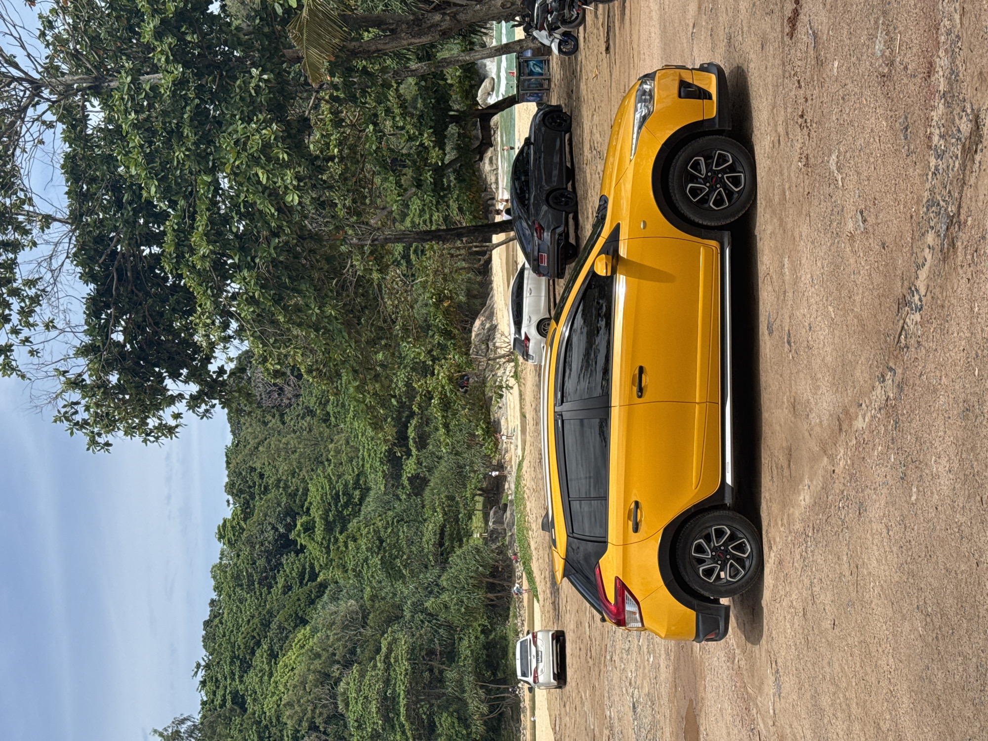 A bright yellow hatchback is parked on a sandy area near a beach in Rawai, Thailand. The car is the main focus, angled towards the right of the frame. Behind the yellow car, several other vehicles are visible, including a white hatchback, a dark sedan, and a motorcycle. The background features a lush, green hillside densely covered with trees, leading down to a sandy beach with gentle waves. Several people are scattered across the beach and near the treeline, some appearing to be walking or standing. The sky is overcast with some patches of blue.