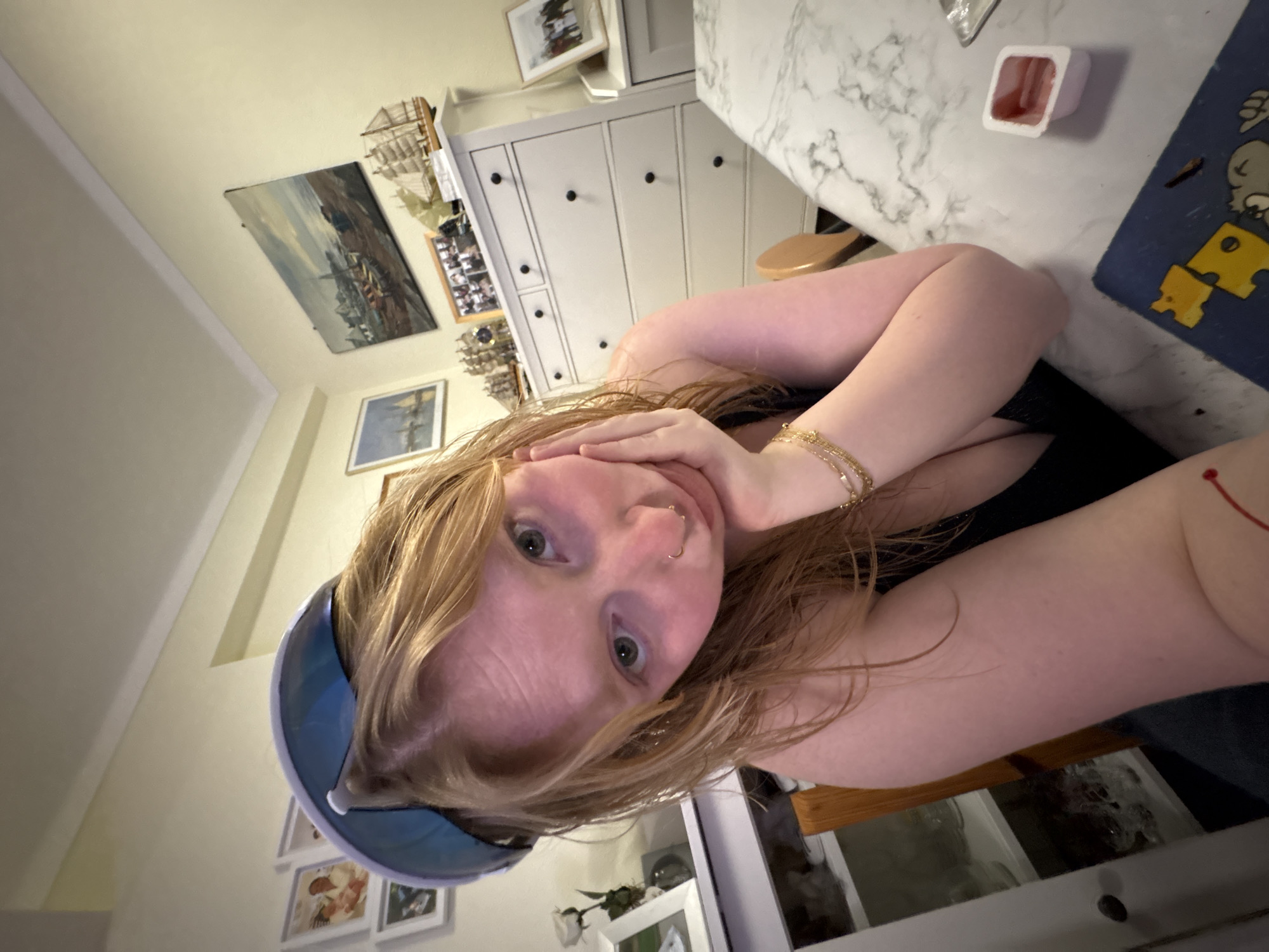 A woman with shoulder-length blonde hair is holding her face in her hands and looking at the camera. She has fair skin, green eyes, and a nose piercing. She is wearing a black top and a blue visor. Her left arm is adorned with gold bracelets, and a red string is visible on her right arm. A white dresser with black knobs stands behind her, accompanied by various framed pictures and decorative items. A white marble countertop is to her right, where a small container with red sauce sits alongside a blue placemat with yellow shapes. The room is illuminated with soft lighting, casting shadows on the wall. The exact city is unknown, but the environment appears to be a domestic setting.