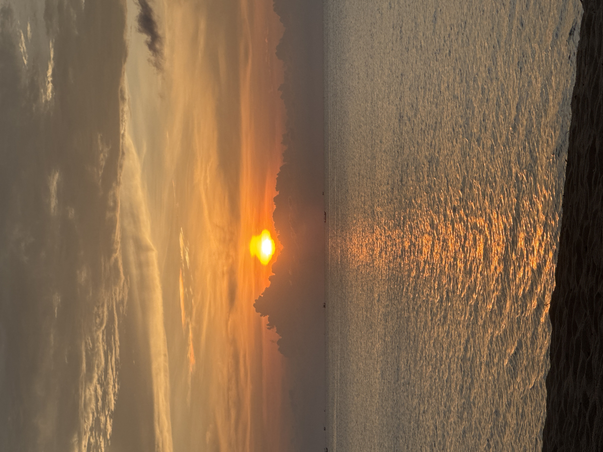 The image captures a sunset over the ocean in Baan Wang Ta Kien, Thailand. The sun is positioned near the center, partially obscured by clouds, creating a warm, orange glow across the sky and reflecting on the water's surface. The sea exhibits rippled texture with light reflecting on the surface. A coastline is visible to the right of the body of water. Small boats dot the horizon. The scene is bathed in a golden light, indicative of the time of day.