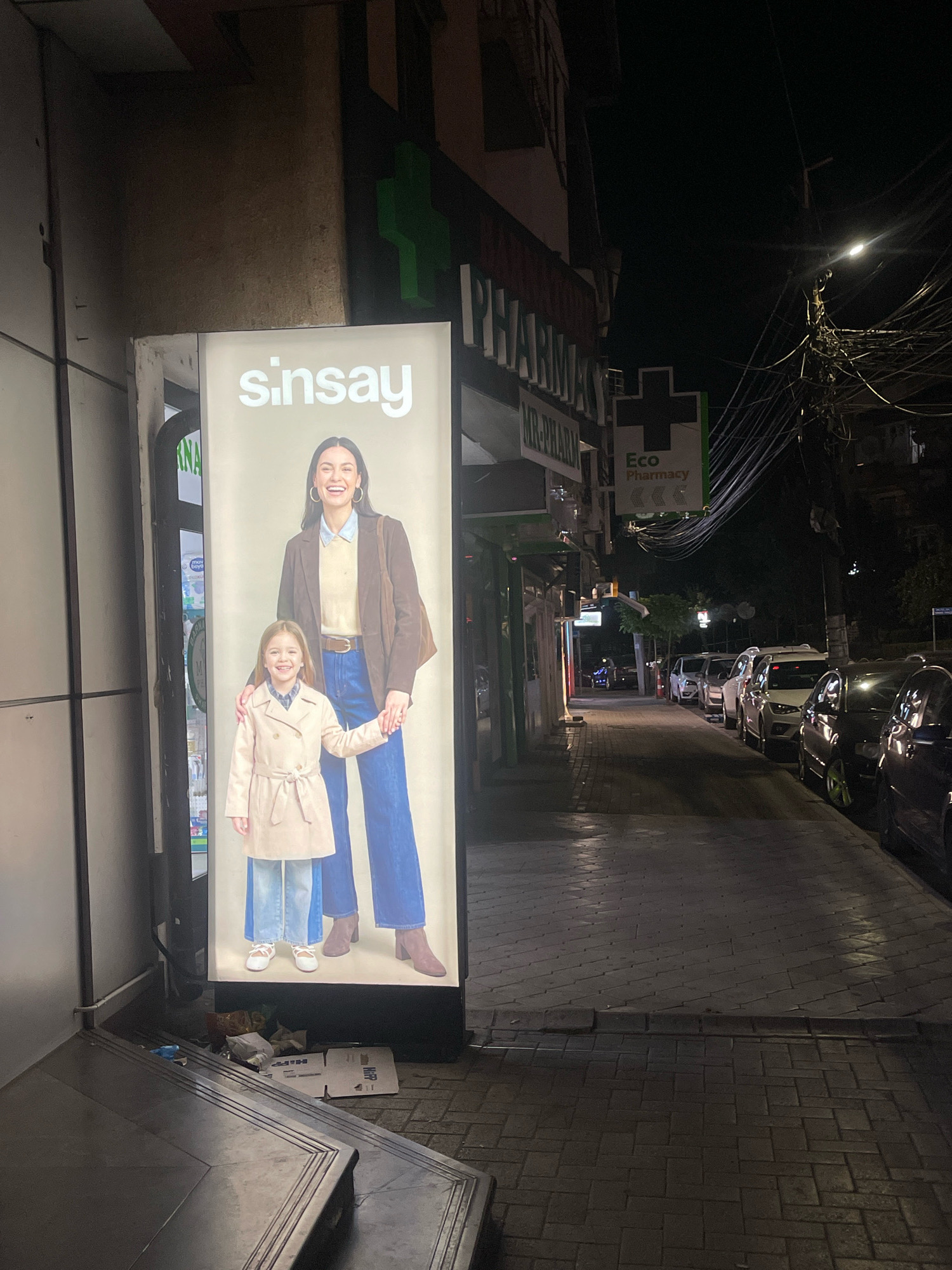 An outdoor, street-level, full-shot photograph, taken at night, shows a lit billboard advertising "Sinsay" clothing on the left side of the frame. The billboard features a woman and a young girl. To the right of the billboard, there are several parked cars, a street, and a sidewalk. In the background, a street lamp illuminates the scene, and there are power lines overhead. A green pharmacy sign is also visible on the building behind the billboard. The area appears to be a commercial street in Prizren, Kosovo. The foreground shows a dark, tiled area with some trash and cardboard boxes.