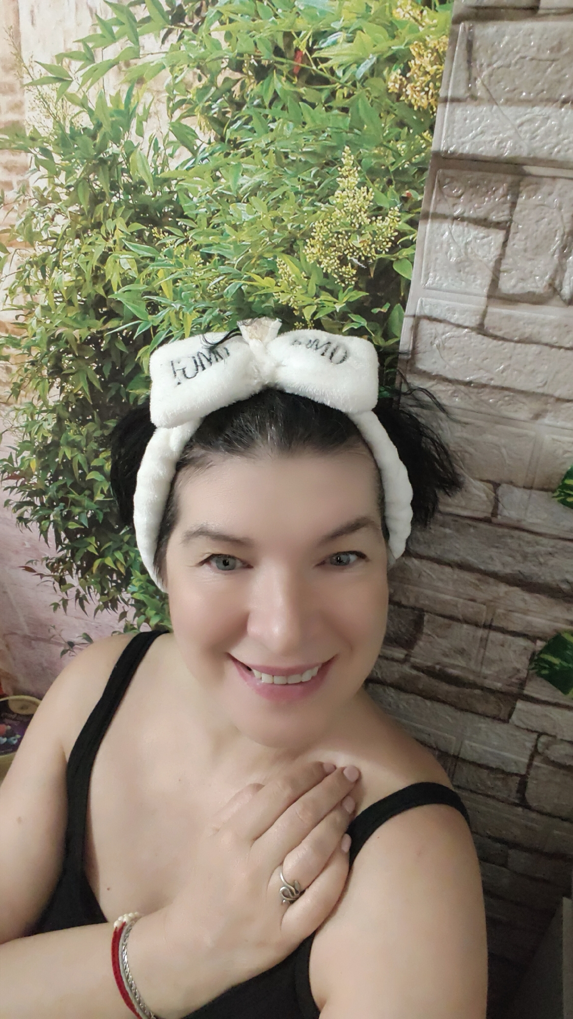 A woman with fair skin and dark hair is smiling at the camera. She is wearing a white headband with a bow on top, and a black tank top. She is wearing a ring on her right hand and has a bracelet on her left wrist. She has her right hand placed on her left shoulder. The background consists of a wall with a faux brick pattern on the right and a backdrop of green foliage on the left.
There is no information to indicate that the photo was taken in the city of Krasnoyarsk, Russia.