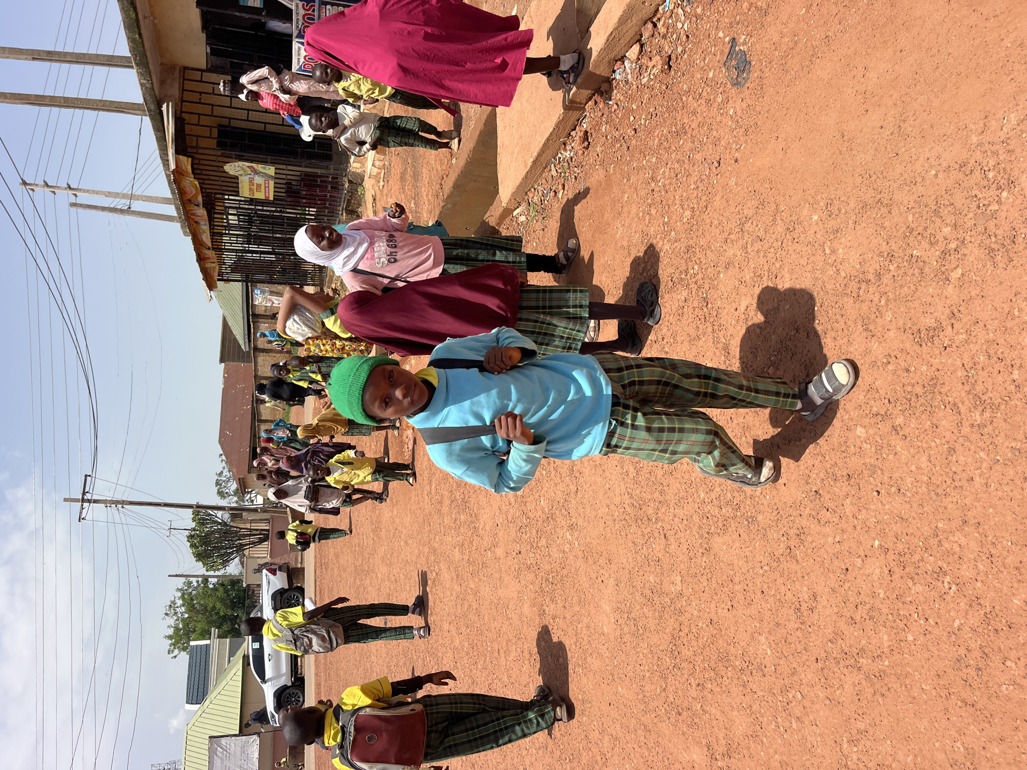 In a dusty outdoor environment with an orange-red dirt ground, several children are walking. The child in the foreground is wearing a bright blue long-sleeved shirt, green and black checkered pants, and a green beanie. This child is also carrying a gray backpack and looking directly at the camera. Behind this child, a group of other children, mostly wearing school uniforms consisting of yellow shirts and green and black checkered pants or skirts, are walking. Some of the children are wearing hijabs or headscarves in various colors including pink and maroon. In the background, there are buildings, including a store with a sign that reads "GLORY SUPER STORE", utility poles with wires, and a white truck parked on the left side of the image. The sky is mostly clear with some clouds. The scene appears to be a street or pathway within a town or city.