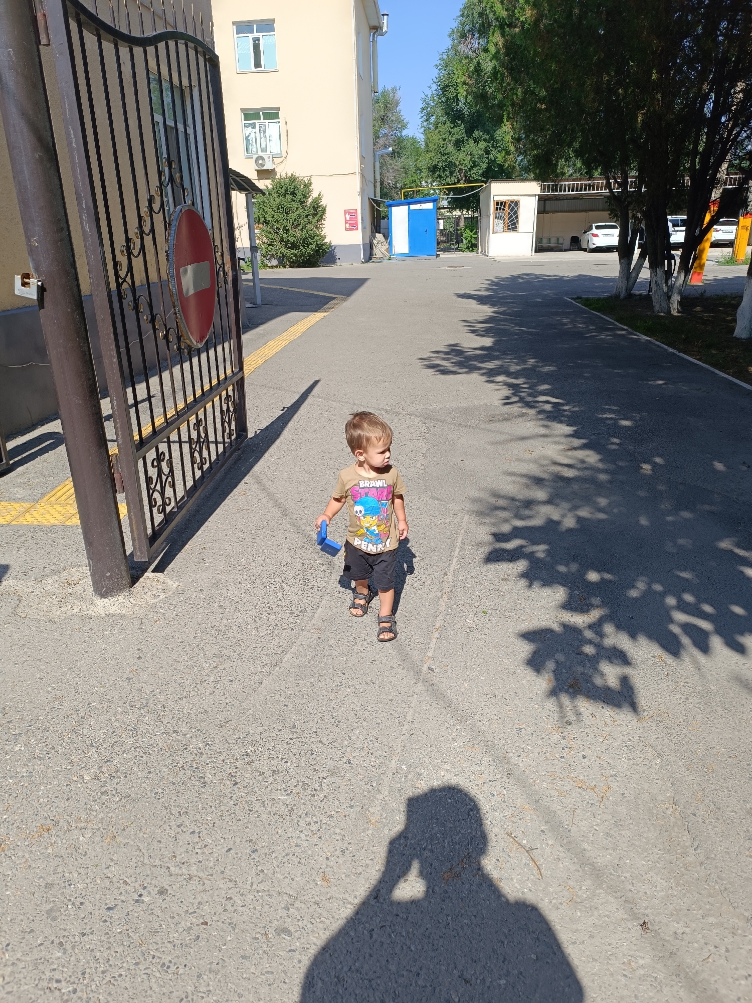 A young boy, approximately three years old, walks on a paved path in Taraz, Kazakhstan. He is wearing a light brown t-shirt with a cartoon character on it and black shorts, along with sandals. The boy is holding a blue toy in his right hand. He is walking away from a metal gate with a red and white circular "no entry" sign. The path is bordered by a yellow tactile paving strip on the left. In the background, there is a light-colored building with windows, trees, and parked cars. Sunlight casts long shadows across the path, including a prominent shadow of the photographer. The environment suggests a sunny day.