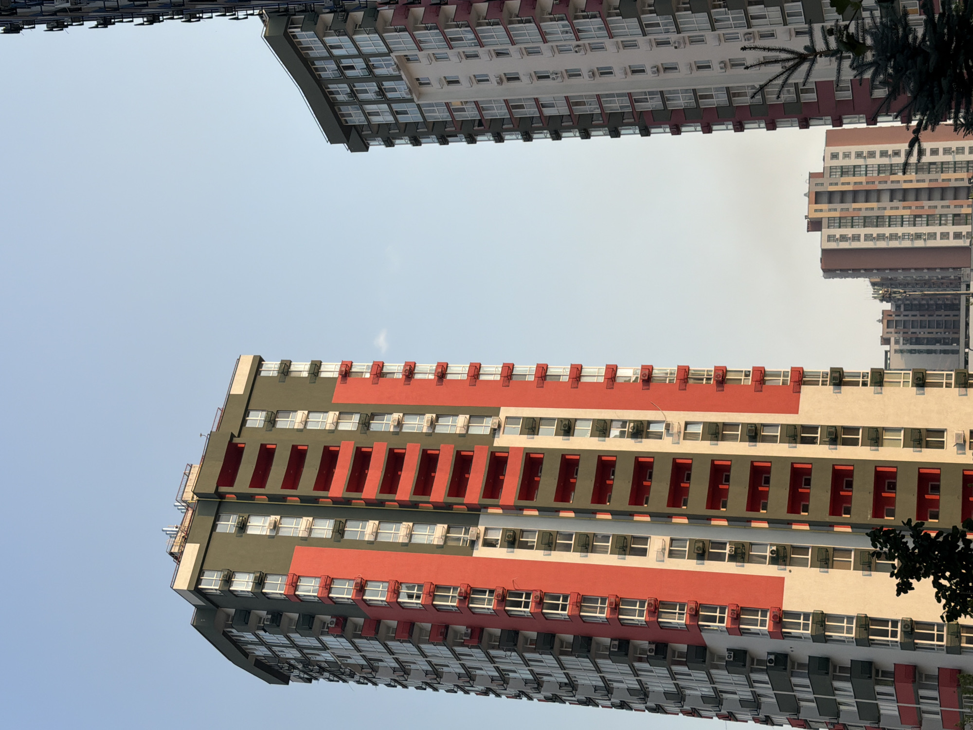 The image shows a clear sky above several modern, high-rise apartment buildings in Kyiv, Ukraine. The building on the left is tall and slender with a striking color scheme of red, grey, and cream. It features numerous balconies and windows, with air conditioning units visible on many of them. The building on the right is also a high-rise, appearing to be slightly shorter than the one on the left, and has a white facade with red accents around the balconies and windows. In the background, a smaller, less colorful apartment building is visible. A small tree is in the foreground on the right side of the image, and a dark tree or shrub is in the lower left corner. The scene appears to be an urban residential area, possibly during daytime with the sun casting shadows.