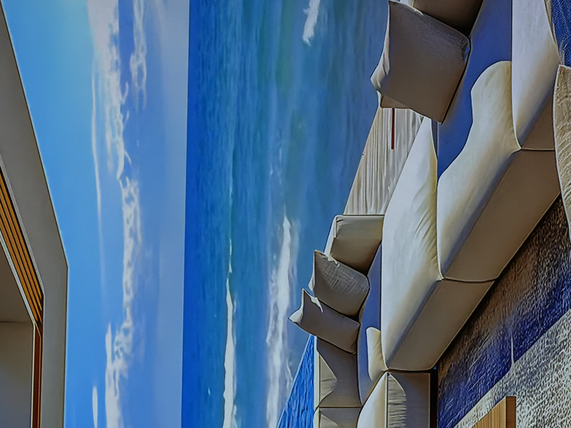 The image captures an upscale indoor lounging area, likely a hotel lobby, showroom, or a high-end residence, within Sofiivska Borshchahivka, Ukraine. The most prominent feature is a massive, seamless digital display or mural occupying the left half of the frame, showcasing a serene and expansive view of a bright blue sky with scattered white clouds merging into a clear blue ocean with gentle waves.
In the right half of the image, an inviting L-shaped outdoor-style sofa is arranged, featuring numerous plush, light-colored (possibly off-white or beige) cushions and throw pillows, hinting at comfort and luxury. The sofa is positioned against a textured wall, which appears to have a gradient of blue and white tones, complementing the digital ocean view. A small section of a contemporary building structure, possibly a wall or archway with horizontal wooden slat detailing, is visible on the far left edge of the scene.
No people are present, lending a tranquil and aspirational atmosphere to the space. The lighting is bright and even, suggesting daytime. The overall impression is one of modern design, luxury, and a clever integration of technology or art to create an immersive, relaxing environment, bringing the feel of a coastal resort indoors. There is no visible text.