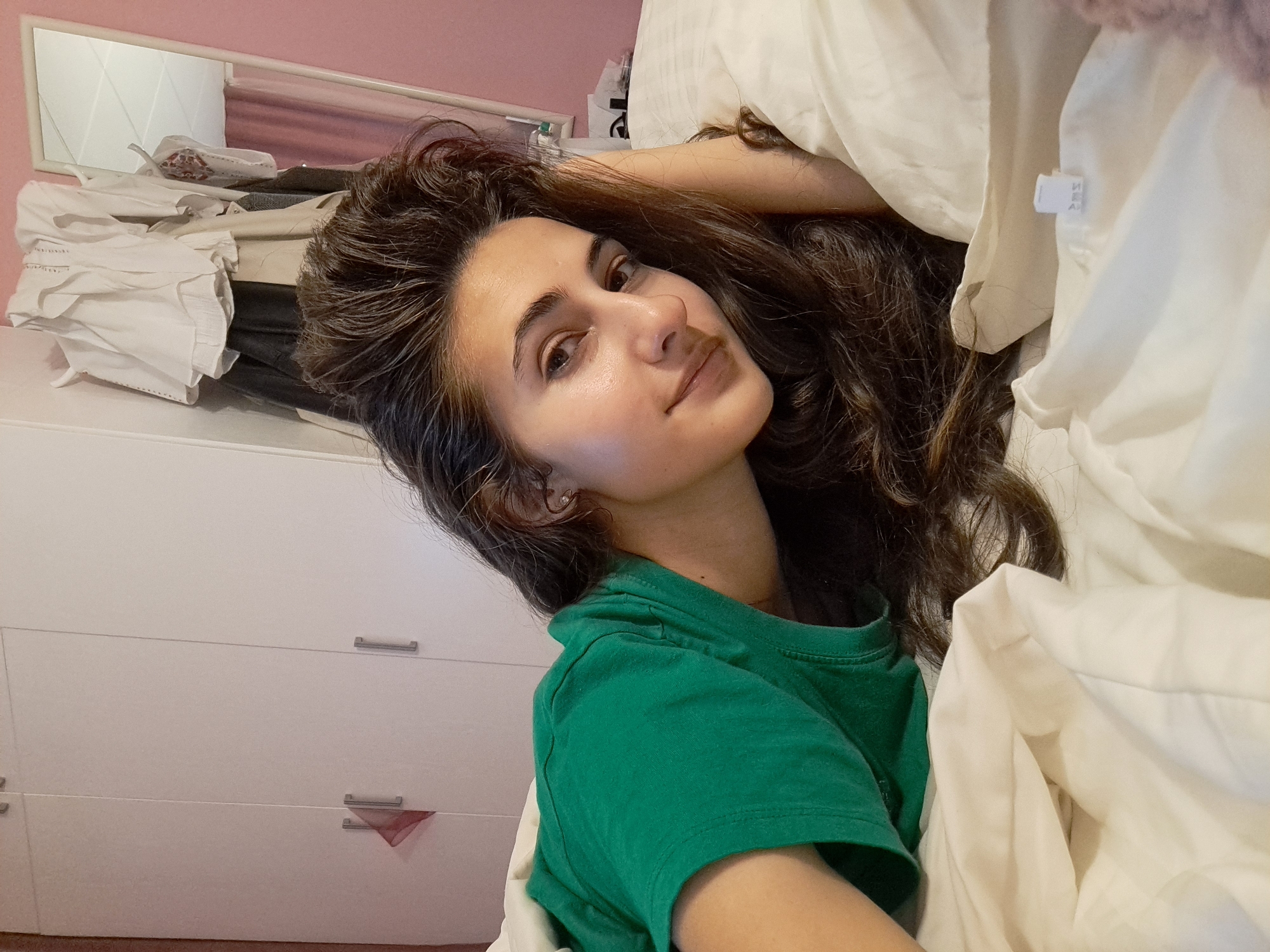 The image depicts a woman with long, dark wavy hair wearing a green t-shirt. She is partially surrounded by white bedding. Behind her is a white dresser with silver handles and a pile of clothes on top. A mirror hangs on a pink wall above the dresser. The location is not discernible as Negovan, Bulgaria, without further visual cues.