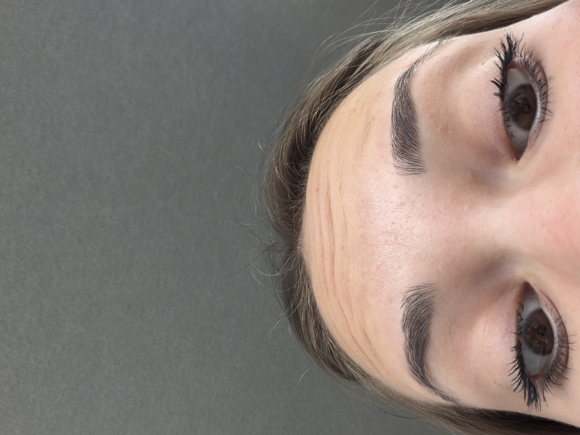The image shows a close-up view of a woman's face, rotated such that the top of her forehead is at the top of the frame. The person has light skin, brown eyes, and meticulously groomed eyebrows. She is wearing mascara. The background is a solid, uniform grey. The city and location are not discernible from the image. There are no actions, interactions, or other objects visible.