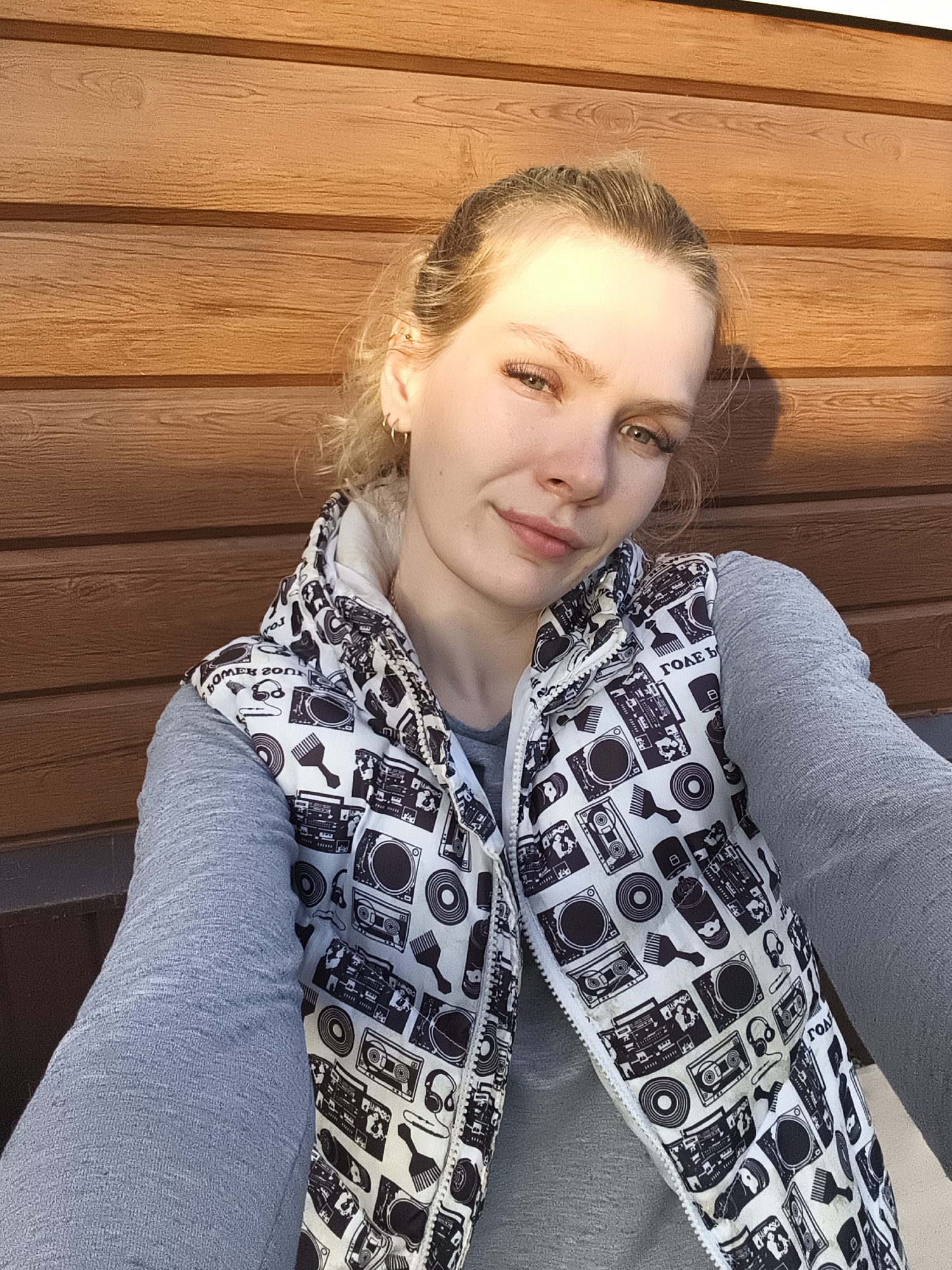 A young woman is posing for a selfie outdoors against a brown wooden wall. She is wearing a grey long-sleeved shirt and a white vest patterned with black musical motifs (boomboxes, records, headphones, etc.). She has blonde hair pulled back, with small gold hoop earrings. The image is taken in bright sunlight. There is no verifiable information about the location within the city of Русский Вожой, Russia, or any other details about specific actions, interactions, or further objects in the environment.