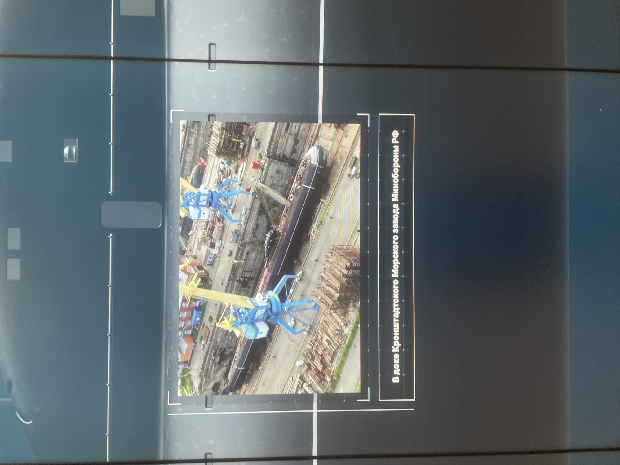 An aerial view shows a large submarine in a dry dock. Two blue and yellow cranes are positioned over the submarine. The dry dock is surrounded by buildings and what appear to be piles of lumber and construction materials. A caption below the image reads "In the dry dock of the Kronstadt Marine Plant of the Ministry of Defense of the Russian Federation."