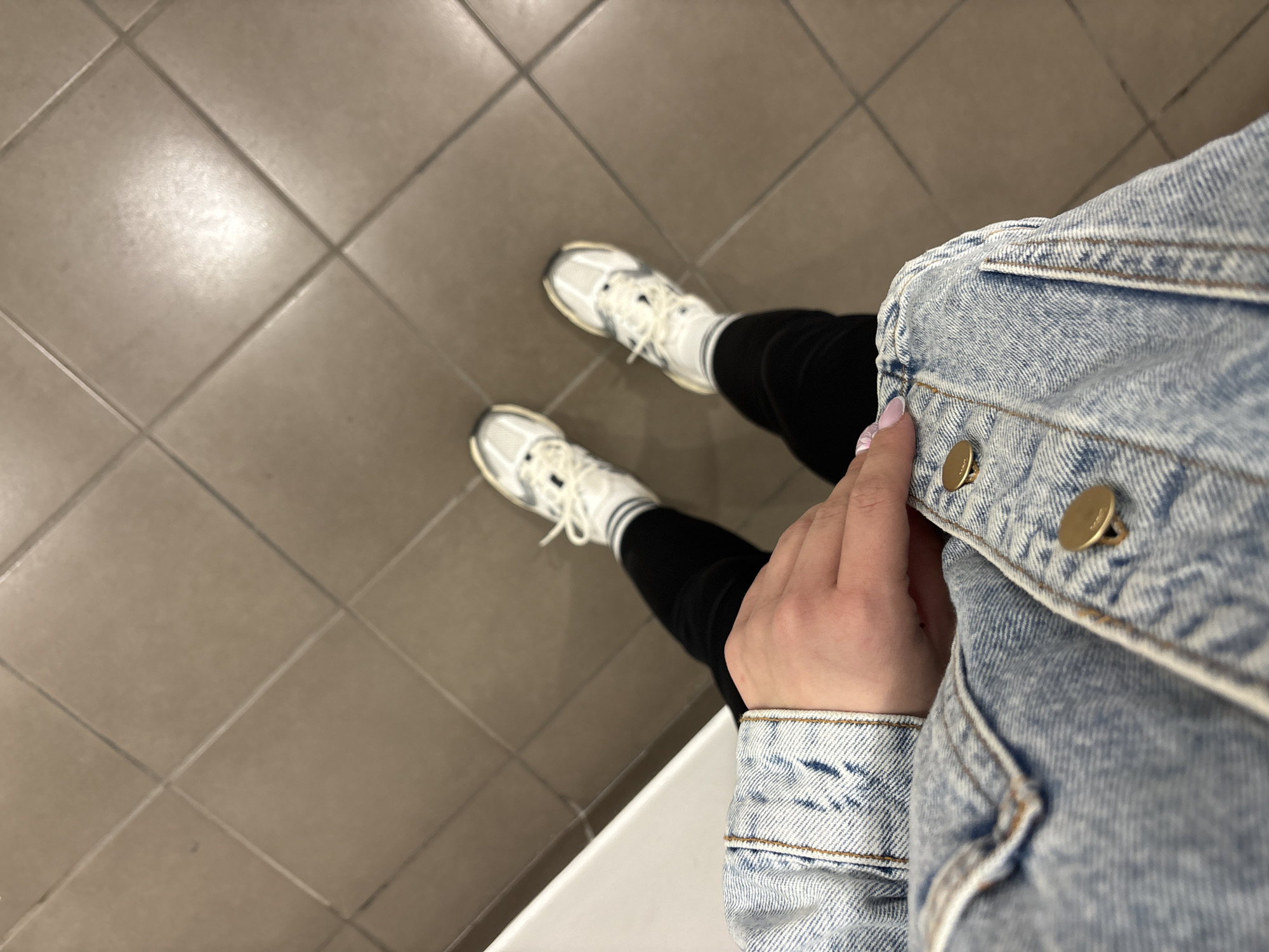 An overhead, point-of-view image captures a person's legs, feet, and a portion of their upper body against a tiled floor, taken in Plovdiv, Bulgaria. The subject is wearing black leggings or pants, white athletic socks with grey stripes at the cuff, and chunky white and grey sneakers. Their left hand, adorned with light pink and white manicured nails, is visible, gently holding the lapel of a light blue denim jacket. The jacket features classic gold-colored buttons and prominent stitching details.
The setting is an indoor space with a beige or light brown tiled floor laid in a grid pattern. A distinct bright reflection on the tiles suggests an overhead artificial light source. In the immediate foreground, a white, possibly ceramic or highly reflective surface is visible, hinting at the person possibly being in a restroom, changing room, or a similar utility area within a public or commercial building. The image depicts a casual, static moment, likely a personal photo taken to show off an outfit or simply capture the surroundings from the individual's perspective. No visible text is present in the image.