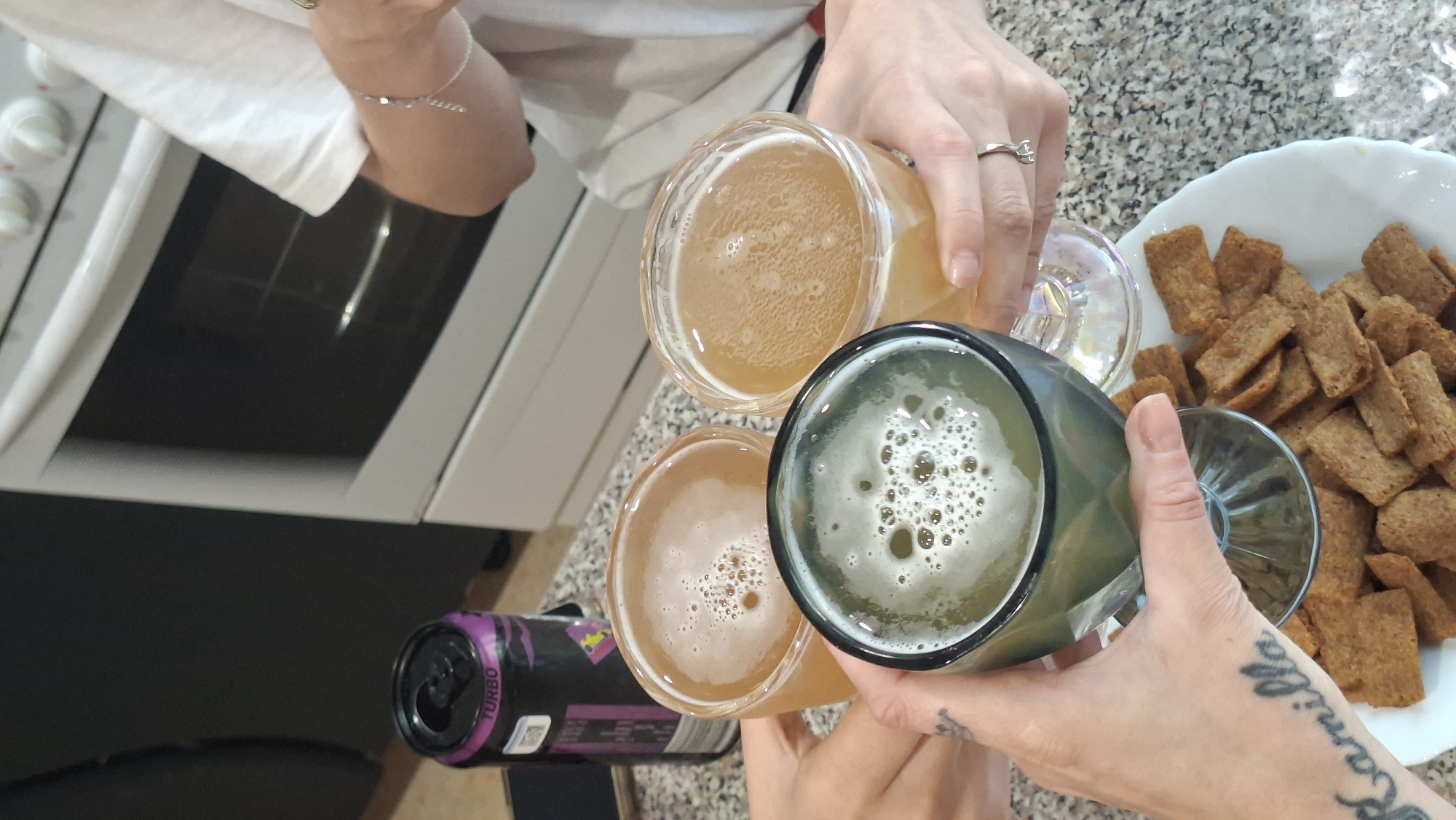 Three glasses of beer are being held up in a toast, with a plate of crackers in the foreground. A black and purple can of "Turbo" energy drink is visible on the left. The scene appears to be in a kitchen, with a white oven in the background. A person's arm with a tattoo of the name "Kamilla" is visible in the lower left corner. The image does not provide any information about the location being Myadzyel, Belarus.