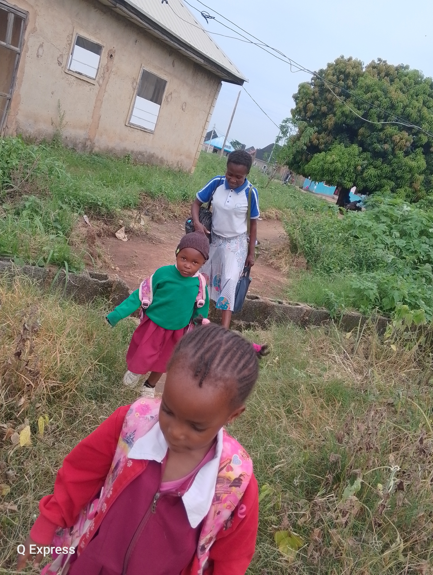In Sabojibi, Nigeria, three individuals are present near a simple, weathered building with two windows. The building appears to be constructed of concrete and has a corrugated metal roof. Surrounding the structure is overgrown vegetation and dry grass, suggesting an unkempt or rural setting.
In the foreground, a young girl is captured from behind, facing away from the camera. She wears a burgundy dress and a bright red jacket adorned with a pink floral pattern.
Behind her, another young girl, looking directly at the camera, is dressed in a green long-sleeved shirt and a burgundy skirt. She has a dark beanie on her head and a pink backpack slung over her shoulders.
Further back, a young woman walks away from the camera, looking over her shoulder and smiling. She is wearing a white polo shirt with blue trim and a patterned skirt. She carries a dark bag in her right hand. The scene suggests the group may be walking or preparing to walk somewhere, possibly to or from school, given the presence of backpacks. The overall environment is modest and suggests a developing area.