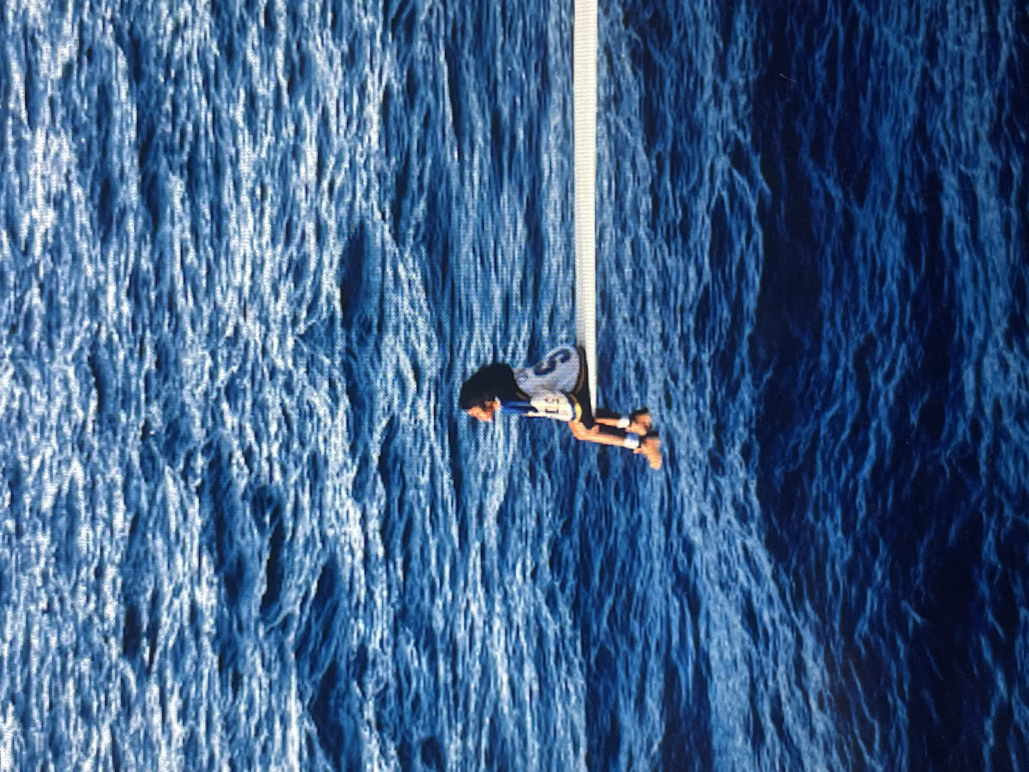 This image captures a high-angle view of a person engaged in a water sport, suspended above a vast expanse of textured blue water. The central subject is a single individual, likely male, clad in dark athletic wear with lighter-colored detailing on their torso and light footwear. They are actively holding onto a prominent, vertical white pole or rope, which extends from the upper part of the frame. The person's body is angled downwards, with bent knees and outstretched arms, indicating active participation in an aerial or towed water activity, such as wakeboarding or a similar cable-supported sport.
The background consists entirely of deep blue water, characterized by its turbulent and rippled surface. Varied shades of blue, from dark to lighter, create a dynamic texture suggesting movement from waves or currents. The perspective is taken from above, looking down on the subject and the water. The lighting indicates daytime conditions, with no harsh shadows, suggesting clear weather. There are no specific landscape features or textual elements visible within the frame. While not evident in the image itself, the provided context places this scene in Sofia, Bulgaria, implying the activity is taking place on a local body of water like a lake or reservoir. The person's posture conveys focus and engagement in the sport.
