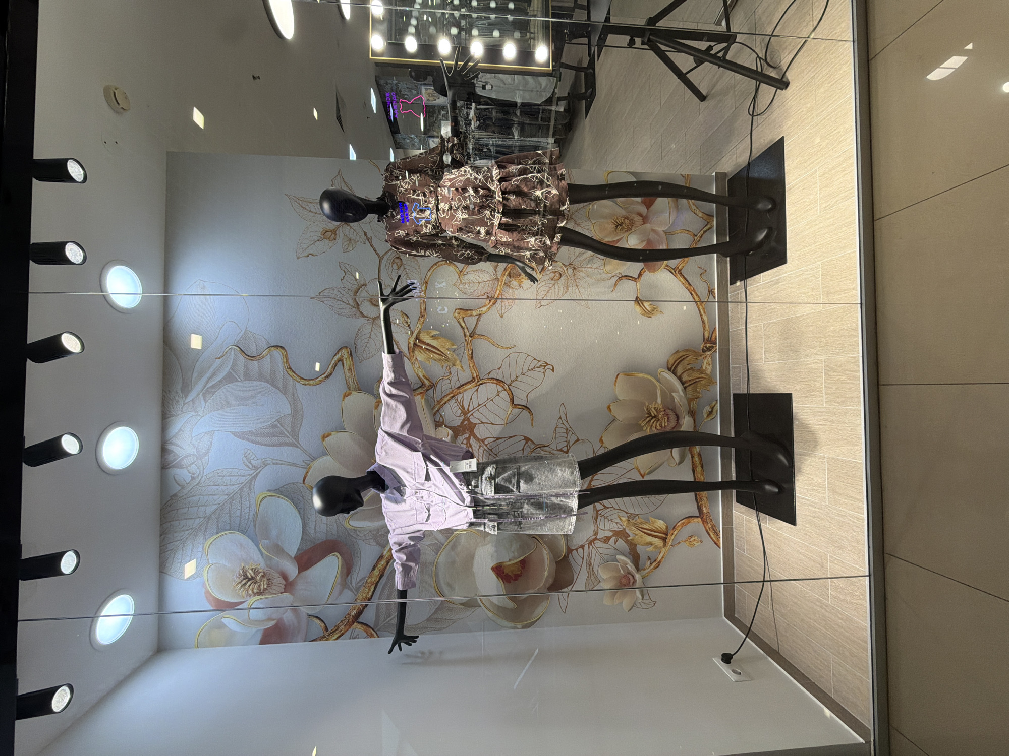 A shop window in Izhevsk, Russia, displays two mannequins against a floral patterned backdrop. The top mannequin wears a brown, patterned dress, while the lower mannequin is dressed in a light purple shirt and silver shorts. The mannequins are black and posed with arms out. The display is illuminated by ceiling spotlights. The window reflects the ceiling and a black metal structure. A power outlet is visible at the bottom of the window. A tiled wall is visible to the right of the window.