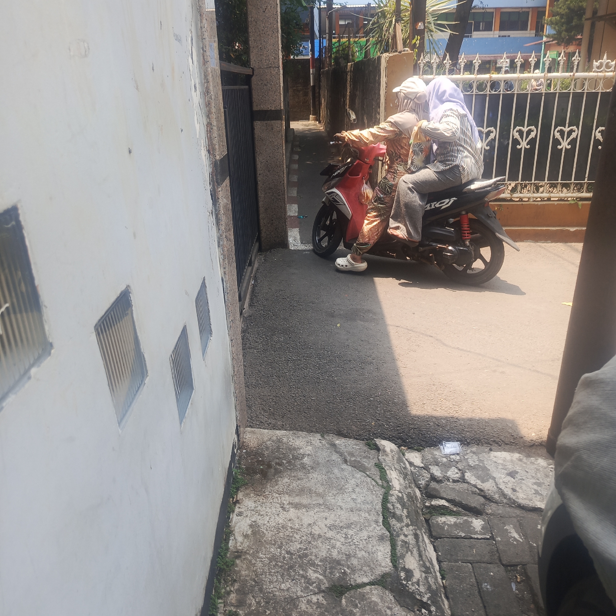 The image depicts a street scene in South Jakarta, Indonesia, with a focus on two individuals riding a red and black scooter. The individuals are wearing modest clothing, with one wearing a light-colored hijab and a patterned top, and the other wearing a hijab and a striped shirt. They are both wearing pants, and one appears to be wearing white clog-style shoes. The scooter is parked on an asphalt road, and a white wall with ventilation blocks is visible to the left. A metal fence with decorative elements runs along the right side of the street, behind the scooter. The environment appears to be a residential or commercial area, with buildings and greenery visible in the background. The lighting suggests it is daytime, with strong sunlight casting shadows. The overall impression is that of an everyday moment captured in a South Jakarta street.