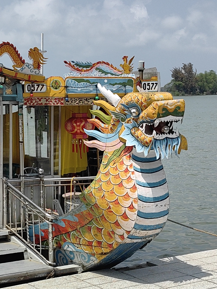 A brightly colored dragon boat is docked on a grey paved surface by a body of water in Thuận Hóa Ward, Vietnam, under a partly cloudy sky. The boat's bow is dominated by a large, ornate dragon head, predominantly yellow and orange, with green, blue, and white accents, featuring white horns and an open mouth exposing teeth. The hull transitions from yellow and orange scales to blue and white horizontal stripes below the dragon head. The upper structure of the boat displays the registration number "0577" on a yellow panel, adjacent to an anchor symbol. This upper deck is adorned with stylized dragons and a phoenix in red, orange, and yellow, set against a blue background depicting bridges and waves. Several red lanterns with yellow tassels hang from the cabin, which has glass windows revealing yellow fabric and a ship's steering wheel inside. Stainless steel railings form an access point from the boat to the dock. The calm body of water extends to the right, with a distant shoreline of dense green trees visible under a hazy sky. Shadows from the boat's structure are cast onto the dock. No people are visible.