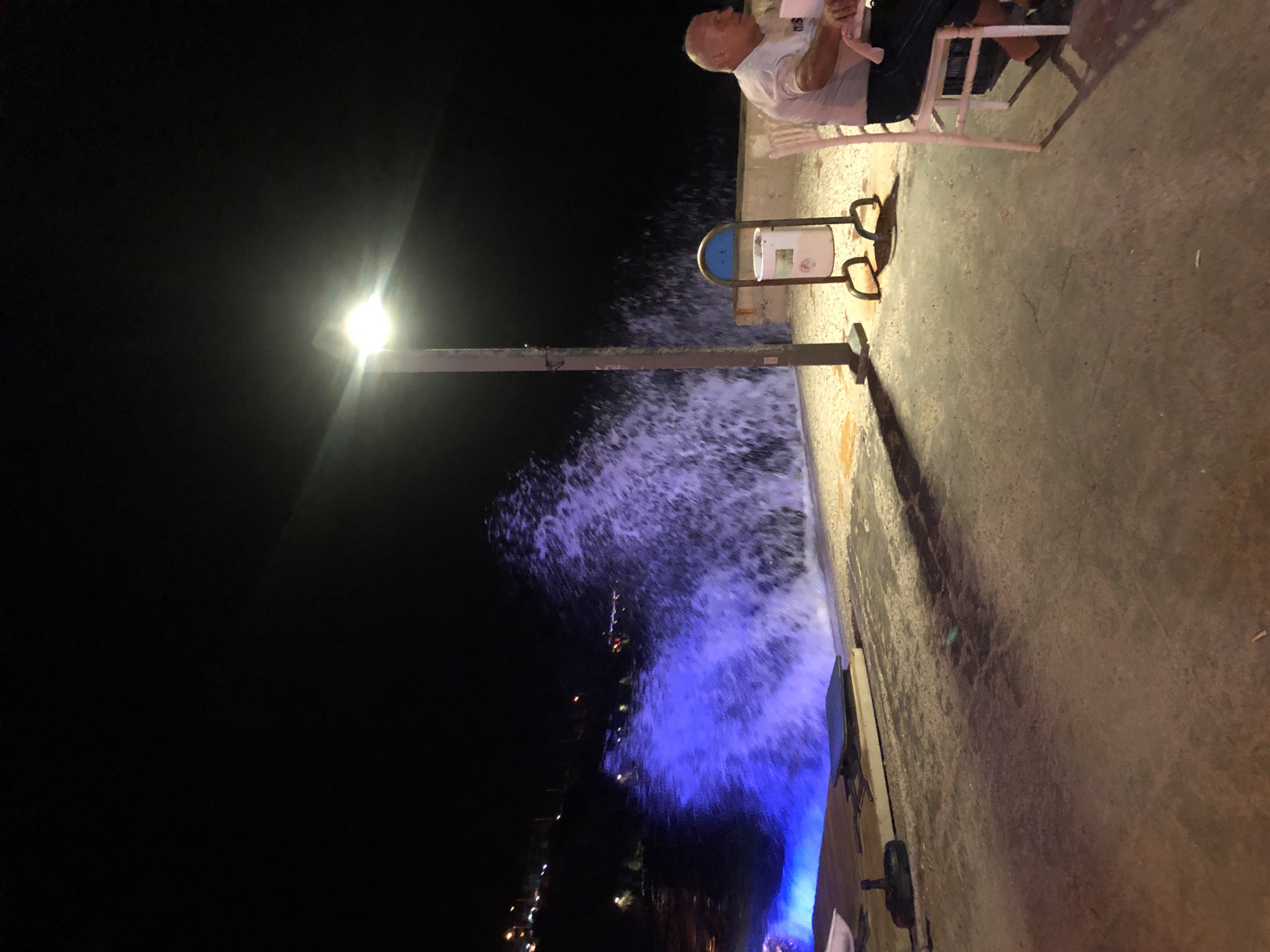 A nighttime scene in Karaburun, Turkey features a large wave crashing against a promenade. The wave is illuminated by a bright blue light, creating a dramatic visual effect. In the foreground, a person sits at a table in a restaurant, seemingly unfazed by the crashing waves. The promenade is lit by a single street lamp, casting long shadows on the ground. The overall atmosphere is one of a lively night out, with the powerful forces of nature providing a dramatic backdrop.