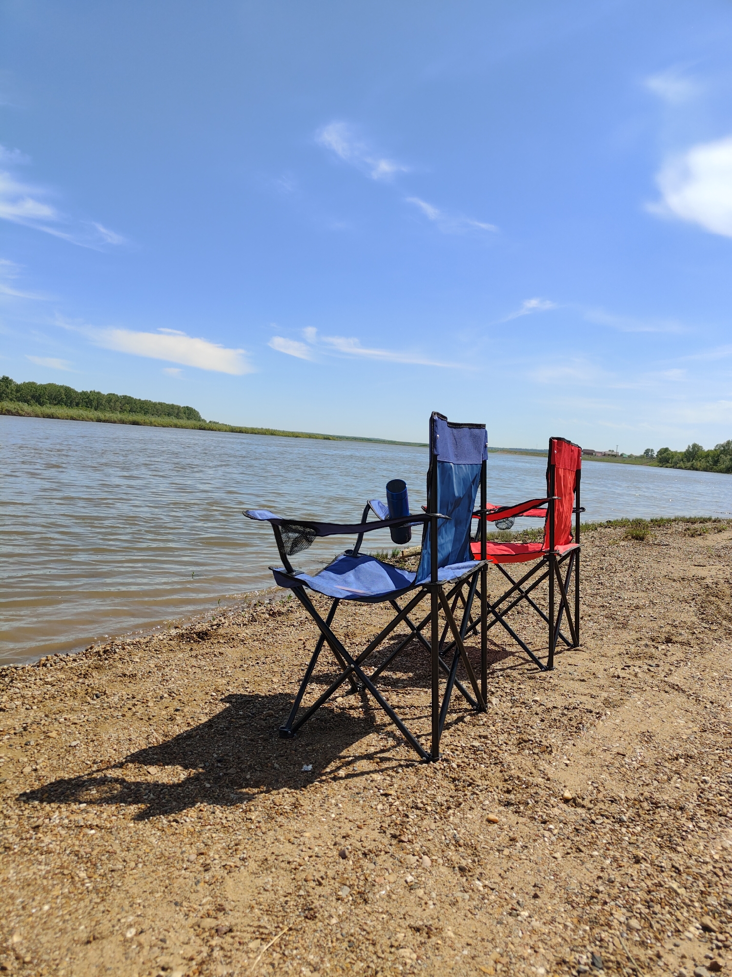 Two folding camp chairs, one blue and one red, stand on a sandy riverbank in Kushnarenkovo, Russia. The river is visible in the background, with a line of green trees on the opposite bank. The sky is blue with scattered white clouds. The chairs are unoccupied.