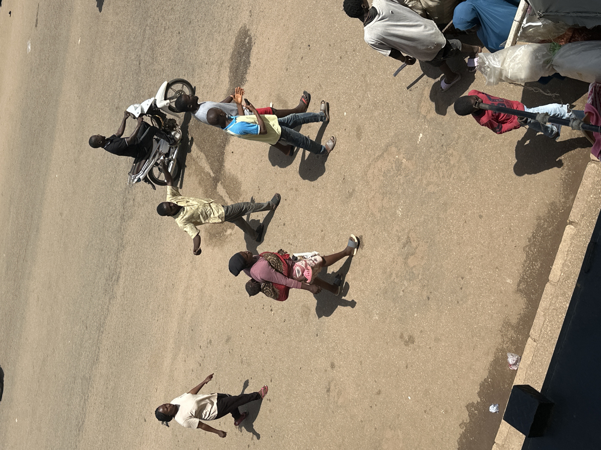 The image depicts an overhead view of a street scene in Bukuru, Nigeria, likely during daylight. Several individuals are visible, some interacting with a motorcycle, others walking or standing.
In the upper left, two men are near a motorcycle. One man in a dark shirt appears to be seated on the motorcycle, while another in a light grey shirt stands nearby. To their right, a man in a blue shirt and jeans is walking, his hands raised. Further down the street, a man in a light yellow shirt and dark pants is standing, looking downwards.
Towards the center right, a woman carrying a child on her back is walking. She is wearing a pink and red patterned outfit and a dark head covering. Another person, wearing a white t-shirt and dark pants, is walking away from the viewer, towards the bottom left. In the upper right, a person in a red shirt and jeans is standing, holding onto a pole. Another individual in a grey shirt is also present in this area.
The ground appears to be a paved street, with some debris and a dark shadow suggesting it is a sunny day. The overall impression is of a normal street scene, possibly a market or a busy thoroughfare. No explicit text is visible in the image.