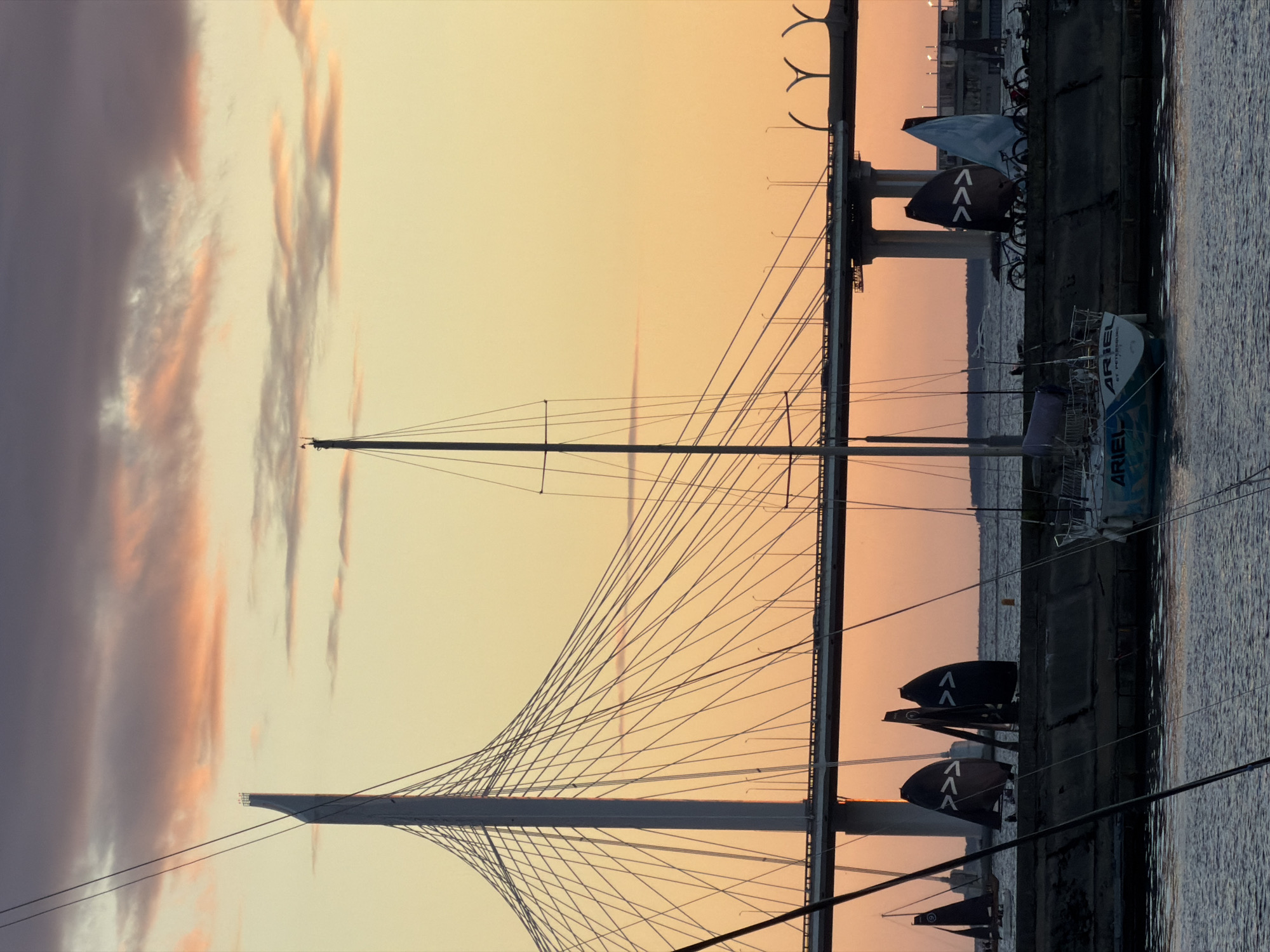 The image captures a serene sunset scene in Saint Petersburg, Russia, featuring a prominent cable-stayed bridge and several sailboats docked along the waterfront. The sky is painted in hues of orange and purple, with wispy clouds adding a touch of drama. In the foreground, the calm water reflects the twilight sky and the silhouettes of the boats.
The bridge, a striking piece of modern architecture, dominates the background. Its white towers and intricate network of cables create a sense of scale and grandeur. Several sailboats, with their masts reaching towards the sky, are moored near the shore. Some have their sails furled, while others appear ready for a regatta.
The waterfront is lined with a concrete embankment, upon which bicycles are parked, suggesting a recreational area or a point of transit. The overall atmosphere is peaceful and contemplative, embodying the beauty of Saint Petersburg at dusk.