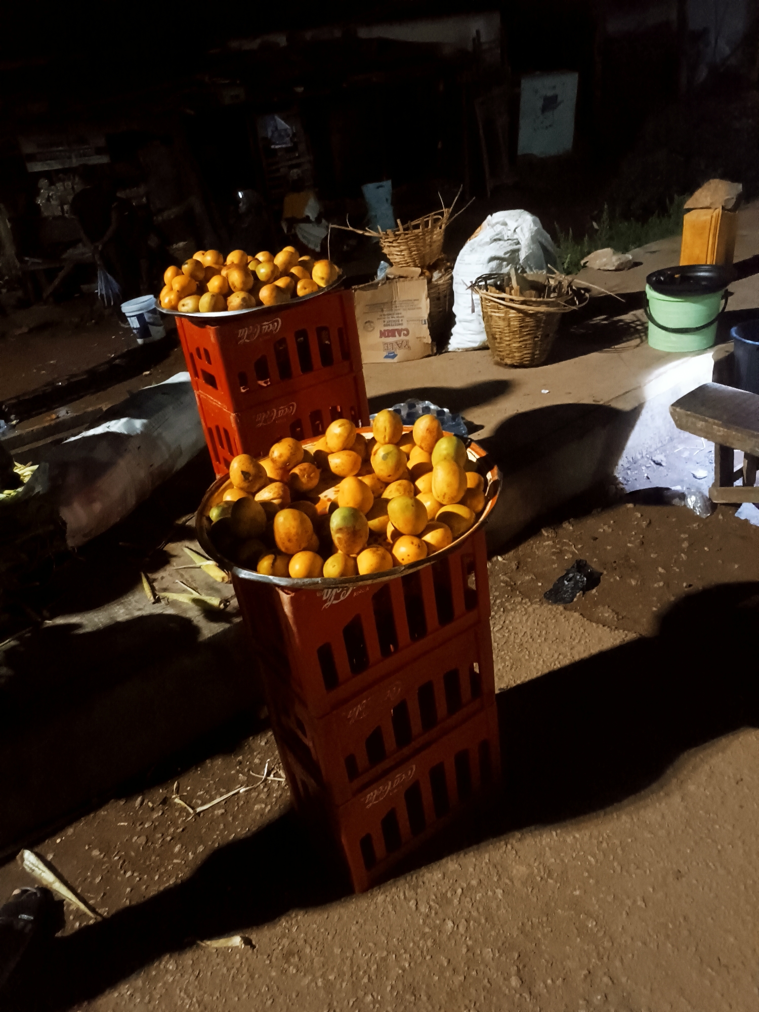 The image, likely taken at night in Bukuru, Nigeria, shows a street scene. Stacks of orange soda crates serve as makeshift stands, each supporting a large bowl filled with mangoes. A small stool and a green bucket are visible near the mangoes. In the background, there are baskets, sacks, and other assorted items. The environment appears to be a roadside market.