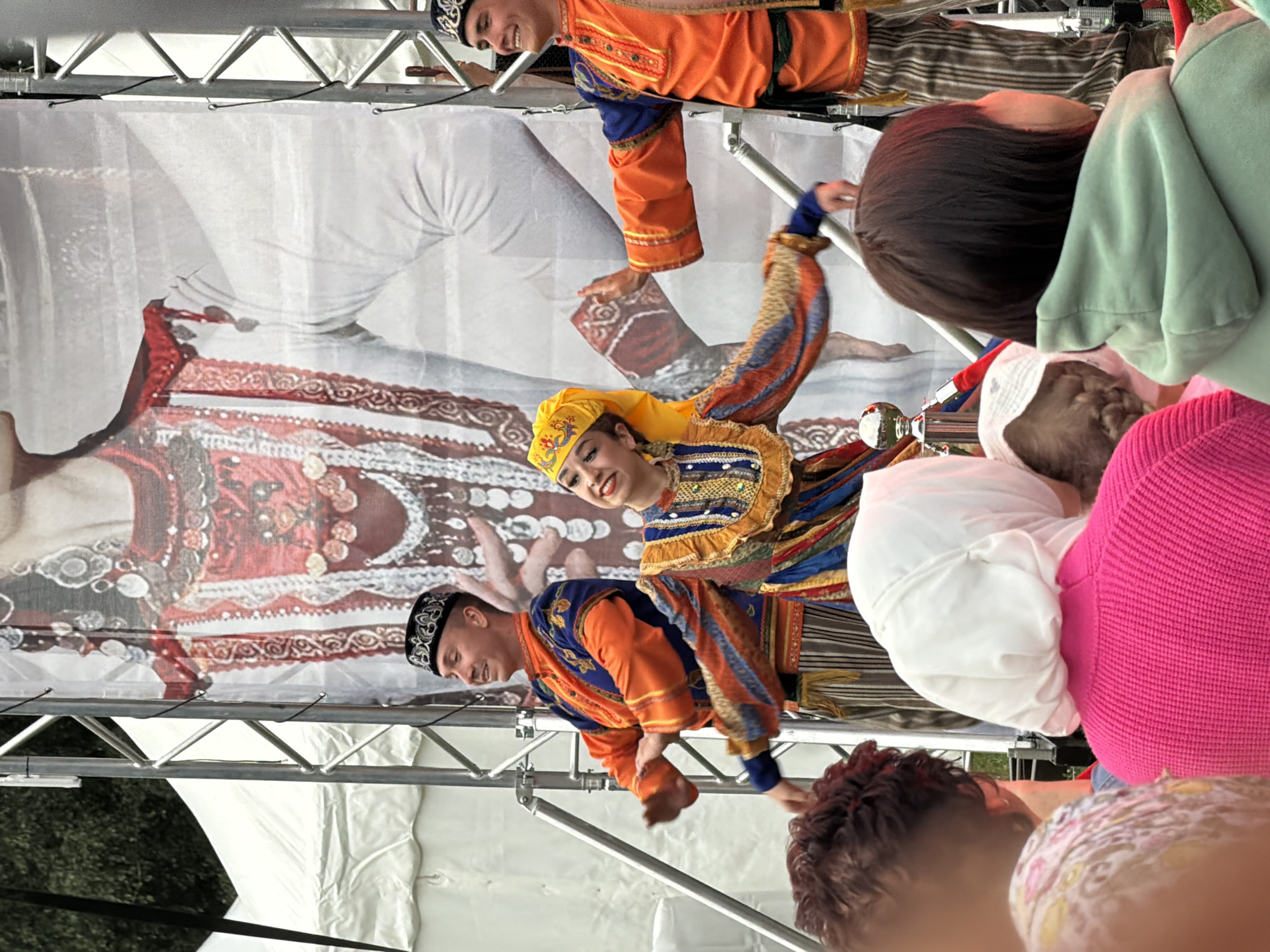 Two male dancers in traditional orange and blue costumes and caps are performing on a stage in Kazan, Russia. A female dancer in a colorful yellow and blue dress is also on stage, dancing with them. In the background, a large banner depicts a person in traditional attire. Several audience members, including a woman in a pink sweater and a woman in a white headscarf, are watching the performance from the foreground. The stage is set up outdoors, with tents and metal structures visible. The event appears to be a cultural performance or festival.