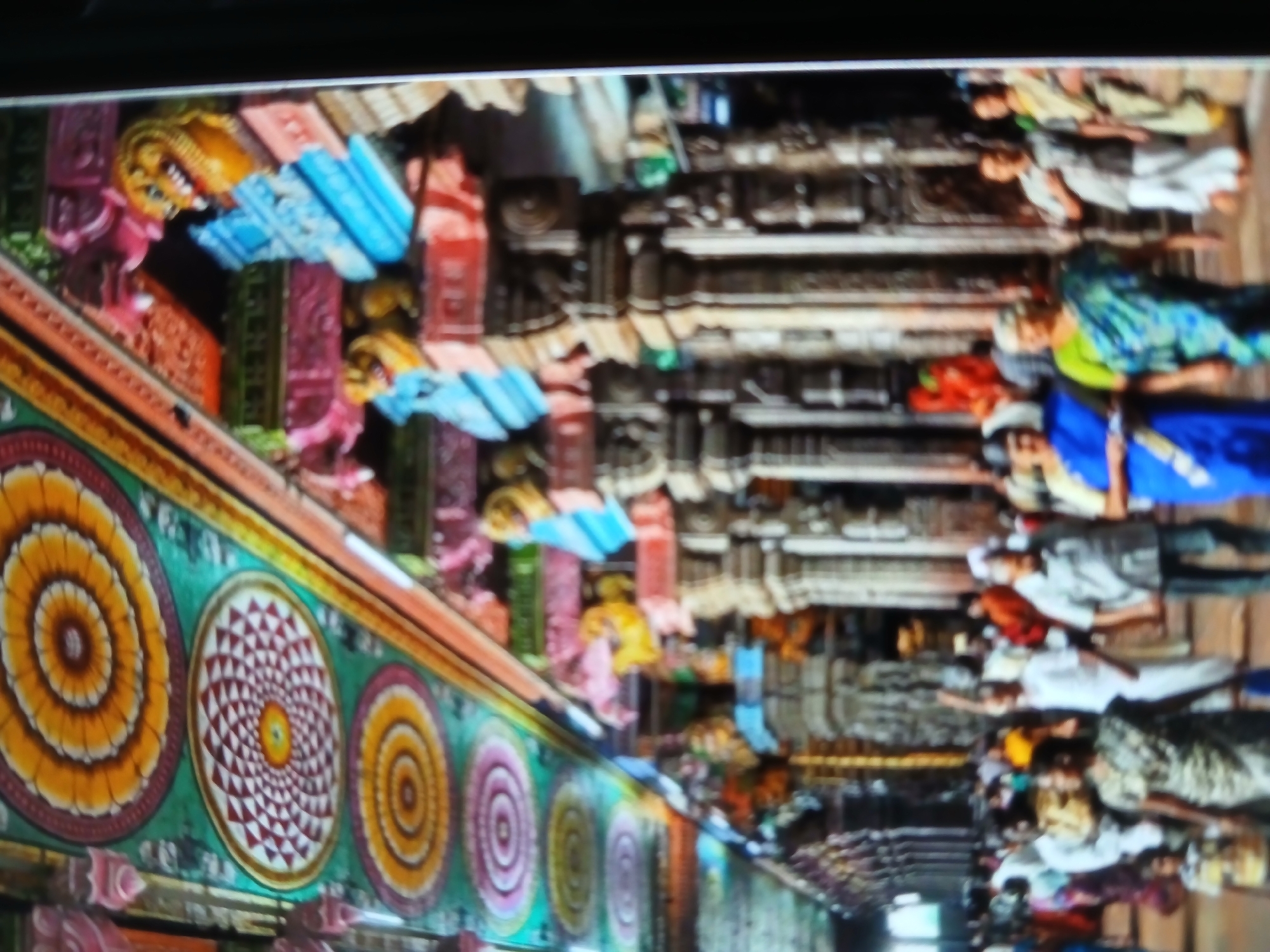 The image shows a crowded street in Bengaluru, India, likely near a temple. The temple's ornate exterior is visible on the left, with colorful circular designs and sculpted figures. A narrow walkway is packed with people, many wearing traditional Indian clothing like saris. The buildings on either side appear to be old and highly decorated. The people are mostly walking or standing, suggesting a busy market or festival.