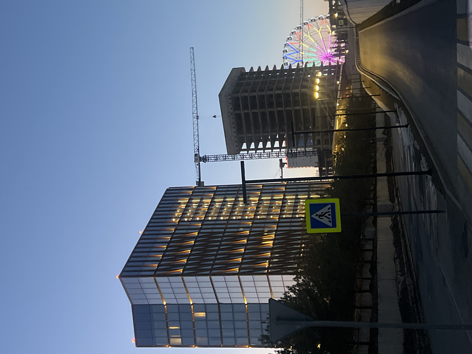 This image shows a street scene in Kazan, Russia at dusk. In the foreground, there is a pedestrian crossing sign and a portion of a street with a zebra crossing. Further back, there is a modern glass building with illuminated internal lights, and a building under construction, including a tall crane. In the background, a brightly lit ferris wheel can be seen. The sky is a gradient of blue to a light orange/yellow.