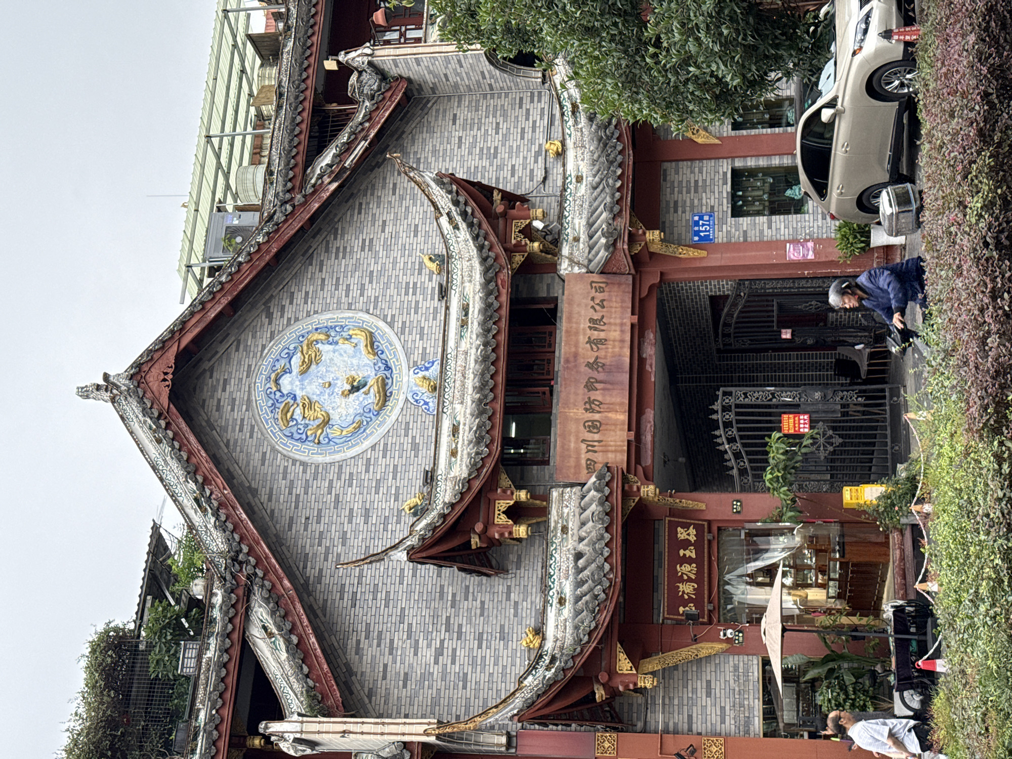 The image captures a street scene featuring a multi-story building with traditional Chinese architectural elements on Citang Street (祠堂街) at address 157 in Qingyang District, Chengdu, China. The building's facade is constructed of light grey bricks, adorned with red wooden structures, tiled roofs with upturned eaves, and gold decorative accents. A large circular blue and gold mosaic depicting two dragons is prominently centered on the upper facade.
Below the dragon mosaic, an arched entrance frames a metal gate, with a weathered brown sign above it displaying Chinese characters reading "四川国防印务有限公司" (Sichuan National Defense Printing Co., Ltd.). To the left of the archway, a ground-floor shop entrance is visible, featuring a dark red facade with gold trim. A sign above this shop reads "玉满源玉翠" (Yuman Yuan Yucui). An open umbrella shades a small outdoor display or seating area near this shop, with potted plants surrounding it.
On the street level, a short, trimmed hedge or planter separates the sidewalk from the building. To the far left, a man in a light-colored shirt is partially visible, standing near a cart with wheels and a red traffic cone. To the right of the building's main archway, a blue street sign clearly shows "祠堂街 157号" (Citang Street No. 157). A red "禁止停车" (No Parking) sign is attached to the metal gate of the main archway. Further right, an elderly person with grey hair, wearing a blue jacket, is seated on and operating a three-wheeled mobility scooter, which has a silver storage container on its rear. Behind the scooter, a gold-colored Lexus RX SUV is parked facing the viewer. The upper sections of the building's roof show various structures, including what appears to be a light green corrugated metal canopy and utility equipment or water tanks, amidst some overgrown greenery. The sky above is overcast and light grey.