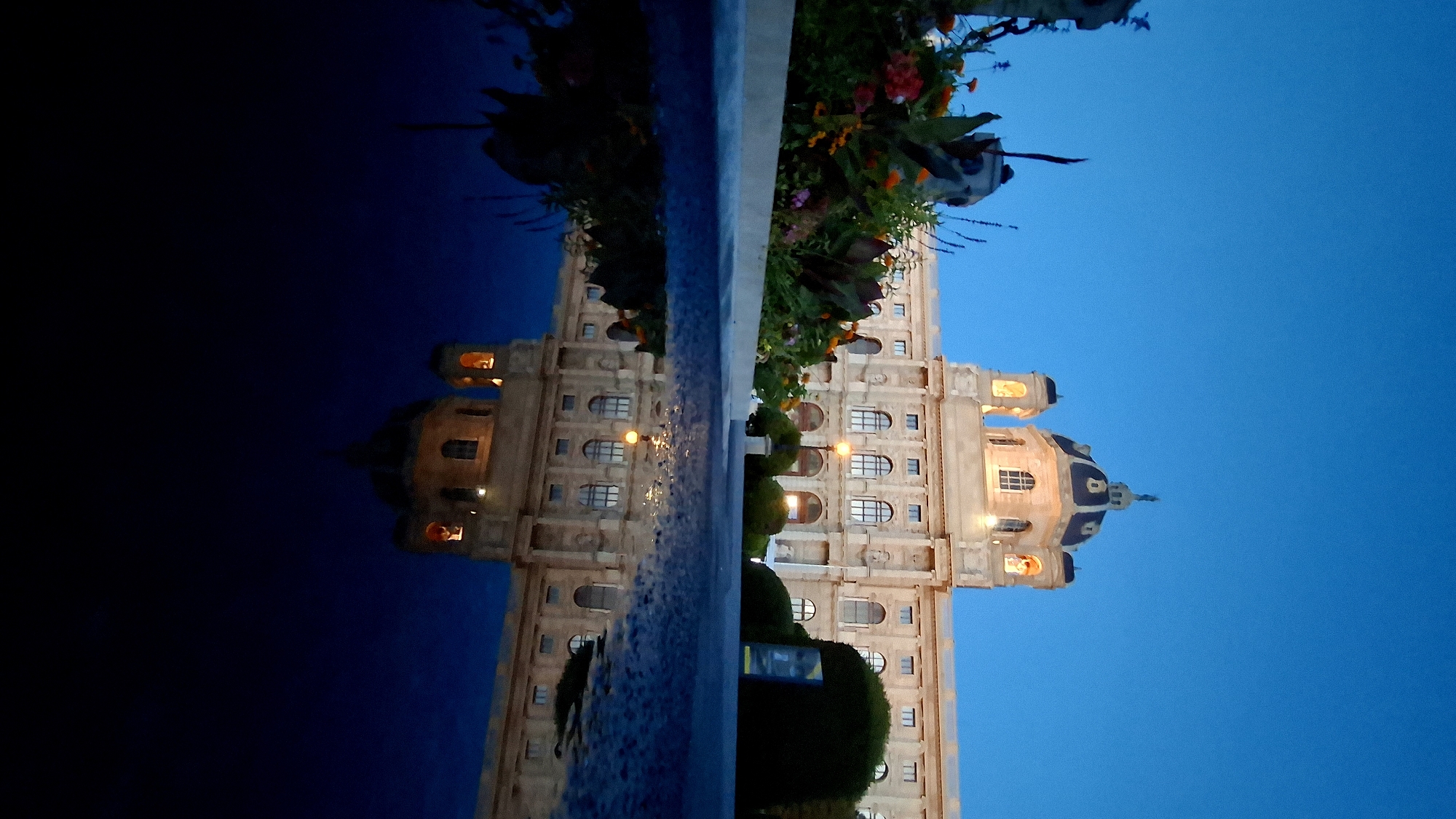 The image captures a low-angle perspective of the Natural History Museum in Vienna, Austria, at dusk. The sky is a deep, clear blue, with the building illuminated by interior lights, highlighting its classical architecture and domed roof. In the foreground, a reflective puddle of water mirrors the museum, creating a striking symmetrical composition. A flower bed with a variety of colorful flowers and greenery is visible to the left of the museum. The reflection in the puddle is darker and less detailed than the actual building, but the distinct dome and windows are clearly visible. The overall scene evokes a sense of tranquility and grandeur.