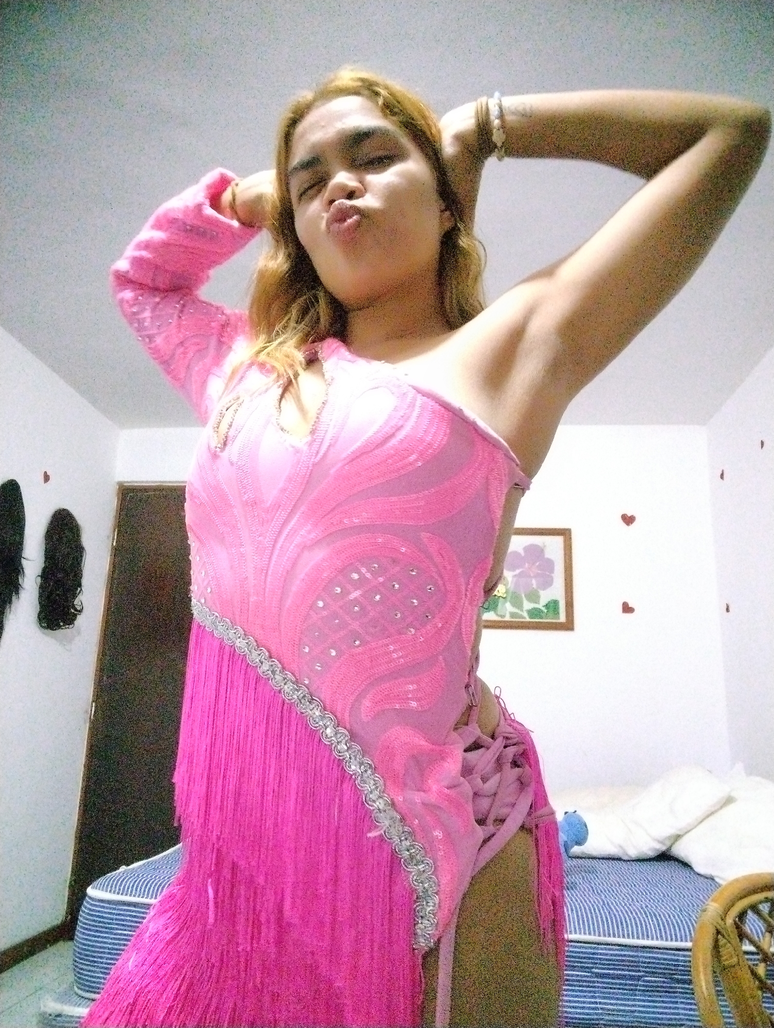 A person, likely female, with light brown hair, is captured indoors posing for a photo. She is making a "kissing" or "pouting" face towards the camera, with both arms raised above her head. She is wearing an elaborate, vibrant pink, one-sleeved costume or dress, featuring sequins, a silver braided trim, and long fringe along the bottom and side. The costume also has a laced-up detail on one side. Bracelets are visible on her right wrist.
The background indicates an indoor setting, possibly a bedroom or dressing room. To the left, a dark brown door is partially visible, and two dark wigs or hair extensions hang on a white wall. Small red heart-shaped stickers adorn the white wall. A framed picture depicting a purple flower hangs on the wall behind the subject's right shoulder. In the lower right portion of the image, a bed with blue and white striped bedding is visible, topped with white pillows and a small light blue plush toy. Part of a wicker chair is also seen in the foreground on the right. The lighting is bright and even, illuminating the scene. There is no visible text in the image.