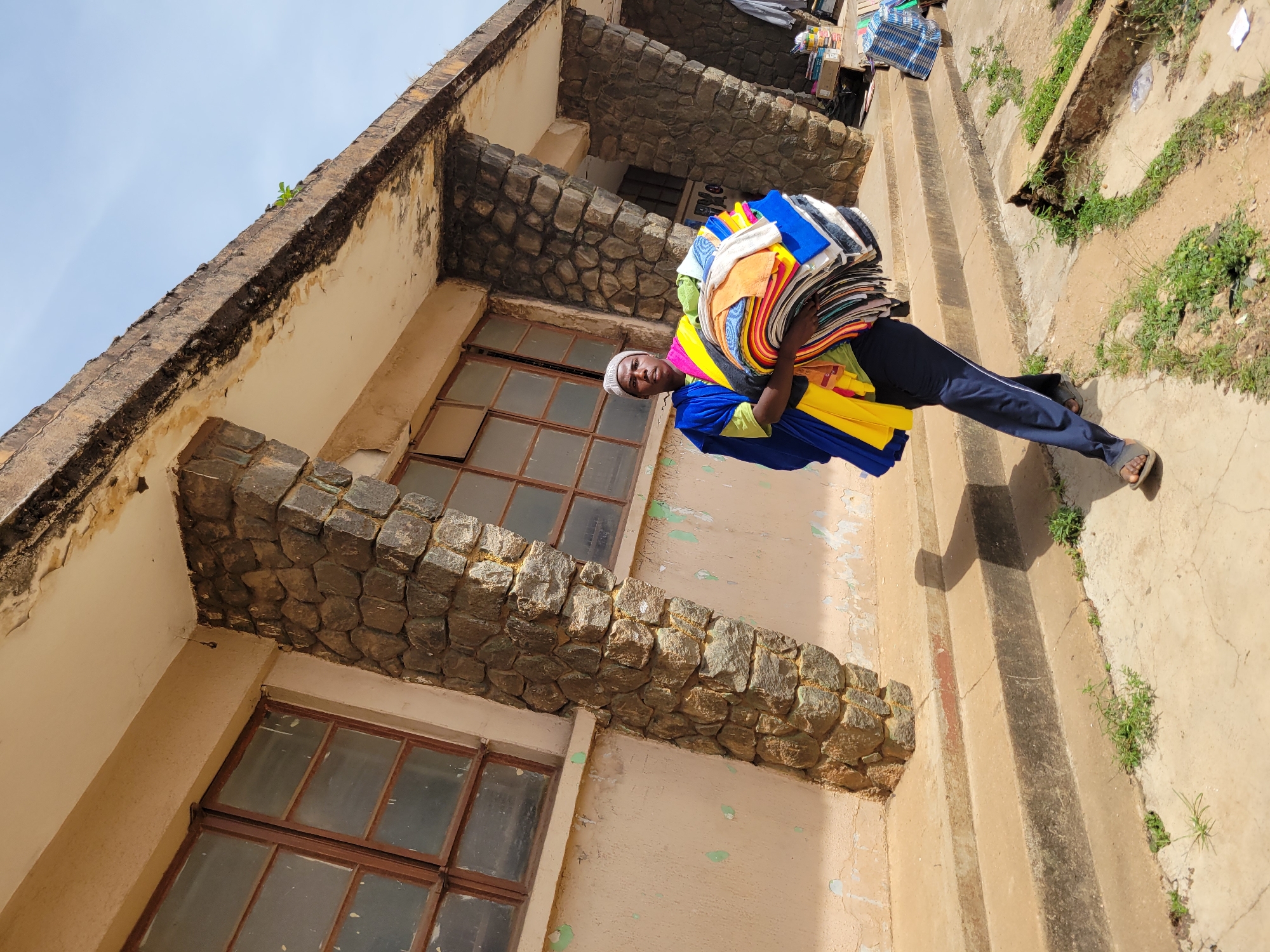 A man carries a large stack of colorful textiles in Jos, Nigeria, standing beside a two-story building with windows framed by brown stone. The building appears to be in disrepair, with peeling paint and discoloration on the walls. The man is wearing a white cap, a blue top, and dark pants. A set of concrete steps and a drainage ditch are visible to the right of the building.