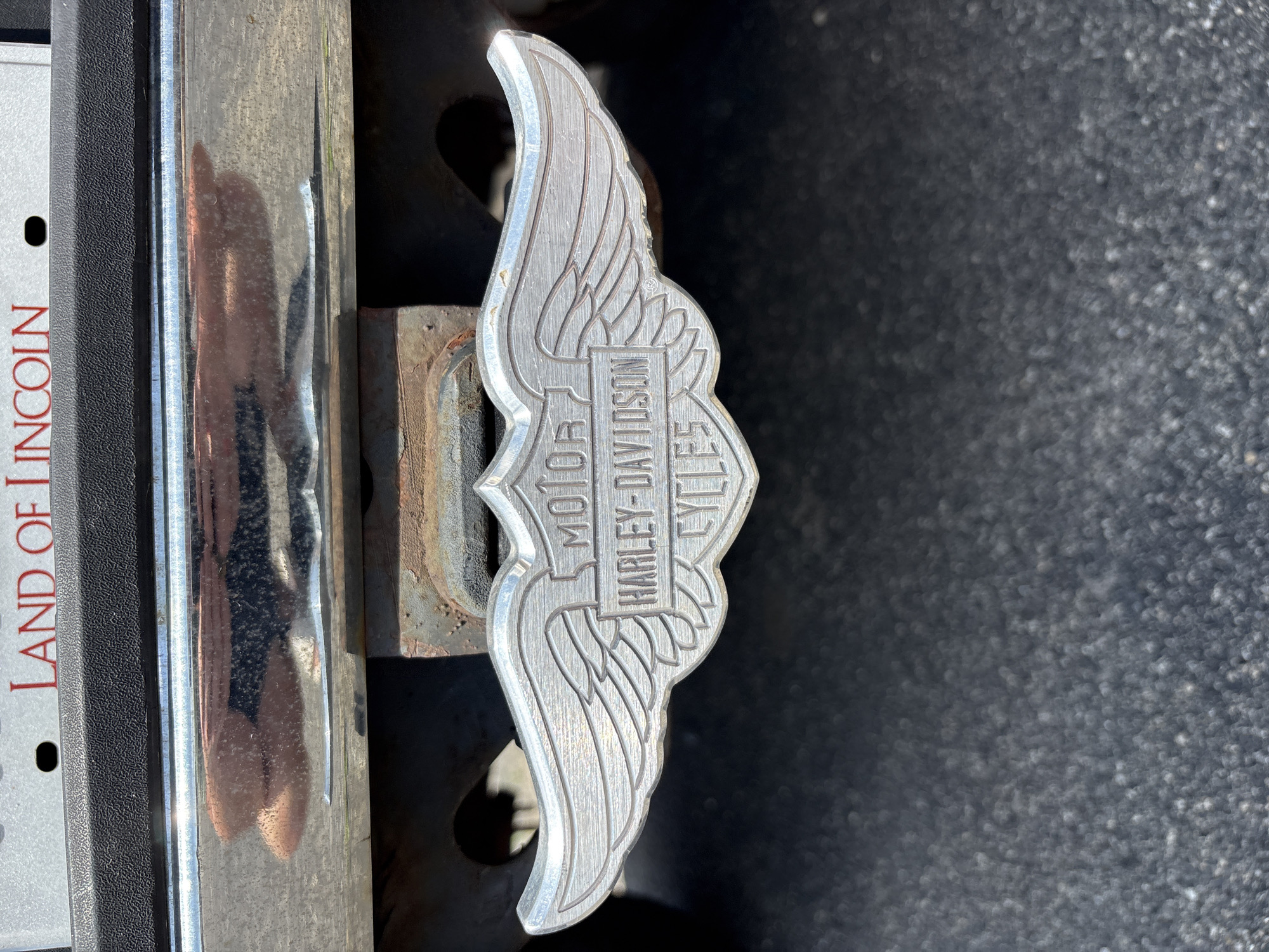 A close-up, eye-level shot shows a silver Harley-Davidson Cycles logo attached to a hitch receiver on the back of a vehicle. The logo is wing-shaped and features the words "MOTOR", "HARLEY-DAVIDSON", and "CYCLES" within a shield design. The hitch receiver is mounted to a dark-colored frame underneath the vehicle's bumper, which is chrome and reflects a blurry image of what appears to be hands and fabric. Above the bumper, a portion of a license plate is visible with the words "LAND OF LINCOLN" in red lettering. The background consists of a dark asphalt surface. The scene is outdoors on a bright day.