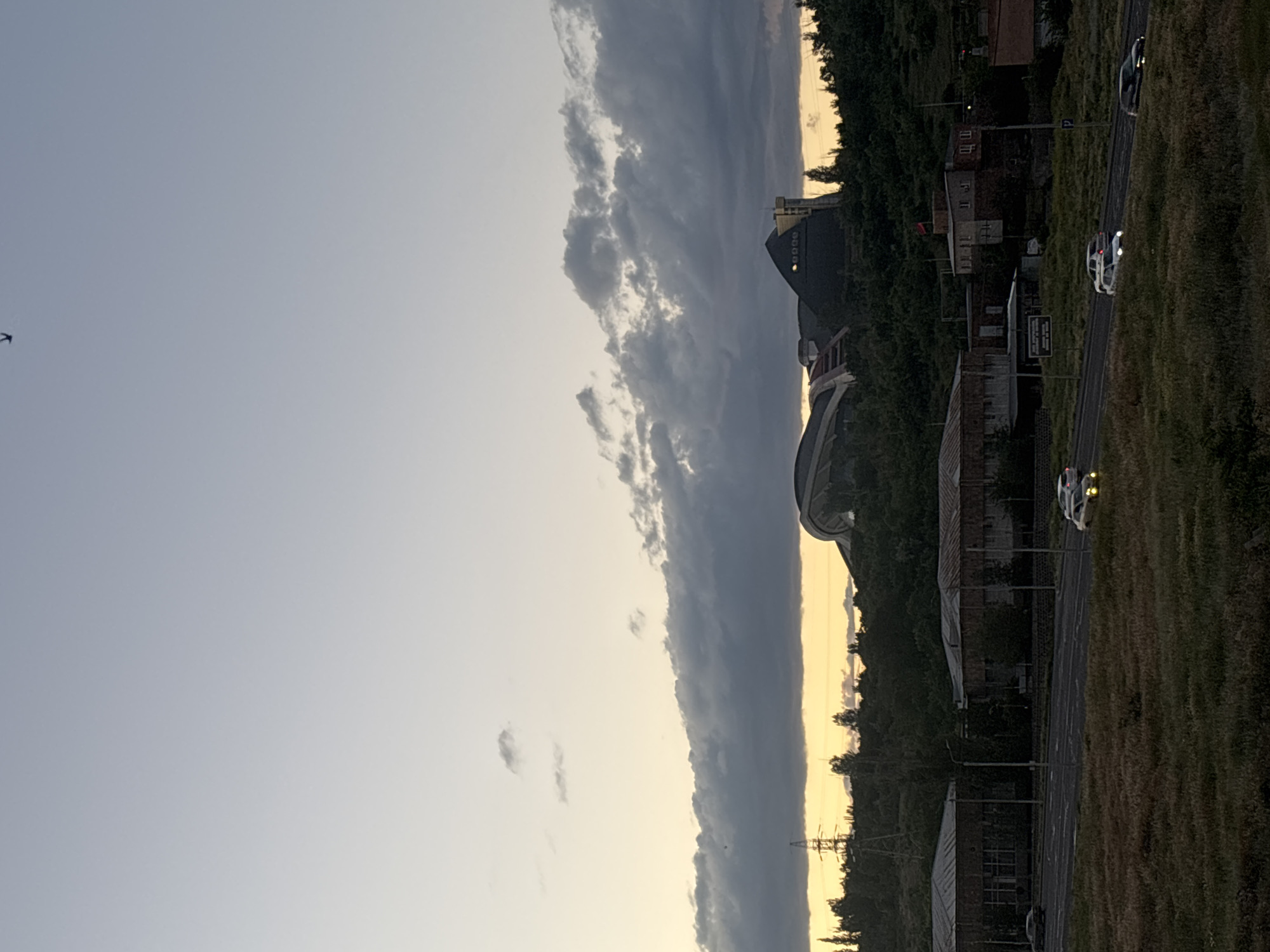 The image captures an urban or suburban landscape at dusk, characterized by transitioning light conditions. The sky occupies the upper portion, featuring a pale blue-grey on the left, giving way to a horizontal band of dark, heavy clouds across the middle, and finally a soft yellowish glow near the horizon on the right, indicative of the setting sun. A small bird is visible in flight in the upper left corner.
Below the sky, the landscape comprises a mixture of natural and man-made elements. A grassy area borders a paved road on the right side of the image. Several vehicles are present on the road, with their headlights illuminated due to the low light. Notably, at least two white cars appear to be police vehicles, identifiable by their design and roof-mounted light bars. A "P" sign for parking is visible near the road.
To the left of the road, a densely treed area obscures some details, but various buildings are visible. These include structures with what appear to be industrial or older architectural styles, alongside more distinct buildings. A prominent feature is a large, dark, dome-like structure, and another building nearby with a distinctive sloped or pointed roof. In the far distance, against the fading light, outlines of hills and power transmission towers can be discerned. The overall atmosphere is calm and quiet, consistent with twilight. No clear legible text is visible on signs or buildings beyond the "P" sign. Given the context, the scene is set in Yerevan, Armenia.