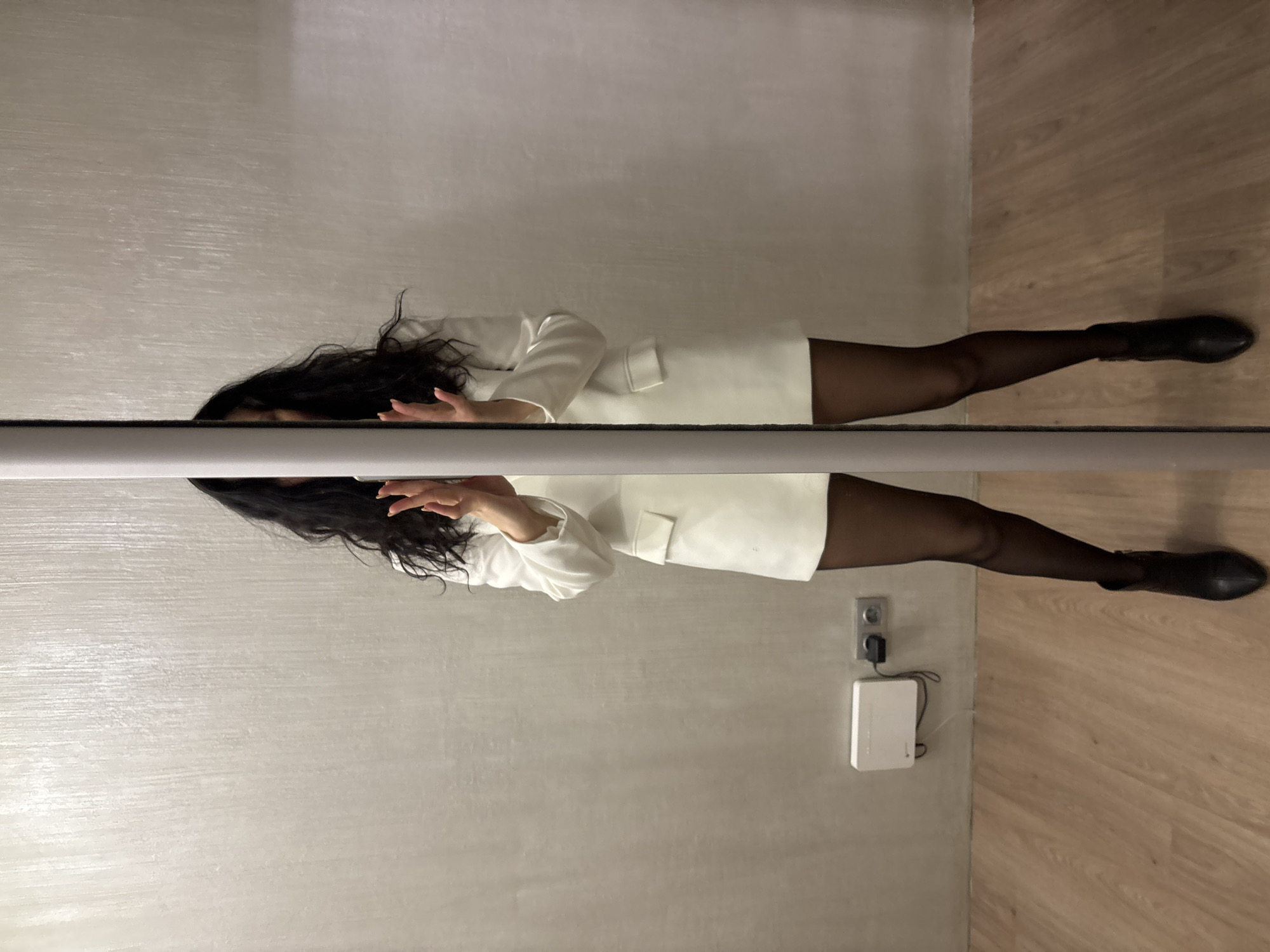 An indoor scene, presumably in Chaikovsky, Russia, features a person captured in a mirror reflection. The original image is rotated 90 degrees clockwise, depicting the person seemingly lying horizontally, with their head towards the left.
The main subject is a person, likely a woman, with long, dark, wavy hair. They are dressed in a stylish white, long-sleeved garment, resembling a blazer-style dress or short coat, paired with dark, opaque tights or stockings and dark, flat-heeled boots. Their face is largely obscured by the edge of the reflective surface in the primary view, but partially visible in the reflection, where they appear to be looking forward. Both hands are slightly raised; in the reflection, one hand shows manicured nails and is held in a relaxed, open gesture.
A prominent horizontal reflective surface, likely a mirror or glass partition, divides the image, creating a clear reflection of the person below. Above this surface, the top half of the person is visible. Below the surface, their reflection is clearly seen, extending down to their feet.
The background behind the person consists of a smooth, light gray or off-white wall with subtle horizontal texture. The floor in front of the person (and beneath the reflective surface in the image) is made of light-toned wooden planks. On the wall, near the bottom right of the reflected image, is a gray wall outlet with a white rectangular electronic device, possibly a modem or smart hub, plugged into it. A small, faint '1' is visible on the device.
The activity appears to be a deliberate pose, possibly for a photograph, with the person observing their reflection. The lighting is uniform, suggesting an indoor setting with artificial light. No specific time of day or strong emotions are discernible.