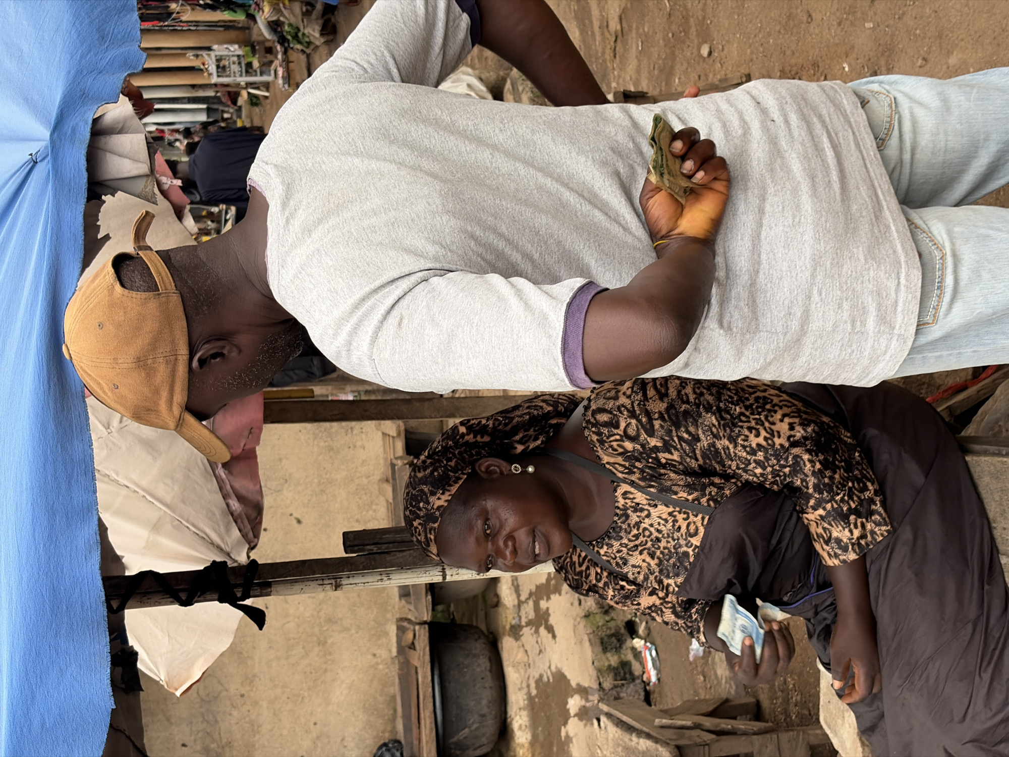 An eye-level medium shot captures two individuals at an outdoor market in Jos, Nigeria. A woman, seated and facing left, wears a patterned headscarf and a dark dress with an animal print overlay. She holds a wad of bills in her right hand. To her right and slightly behind her, a man wearing a light gray t-shirt and a brown baseball cap stands facing away from the camera. His left hand is behind his back, holding a fan of bills. The background shows a makeshift market stall with a blue tarp overhead and various goods and structures typical of a Nigerian marketplace. The ground is dirt and a few discarded items are visible.
