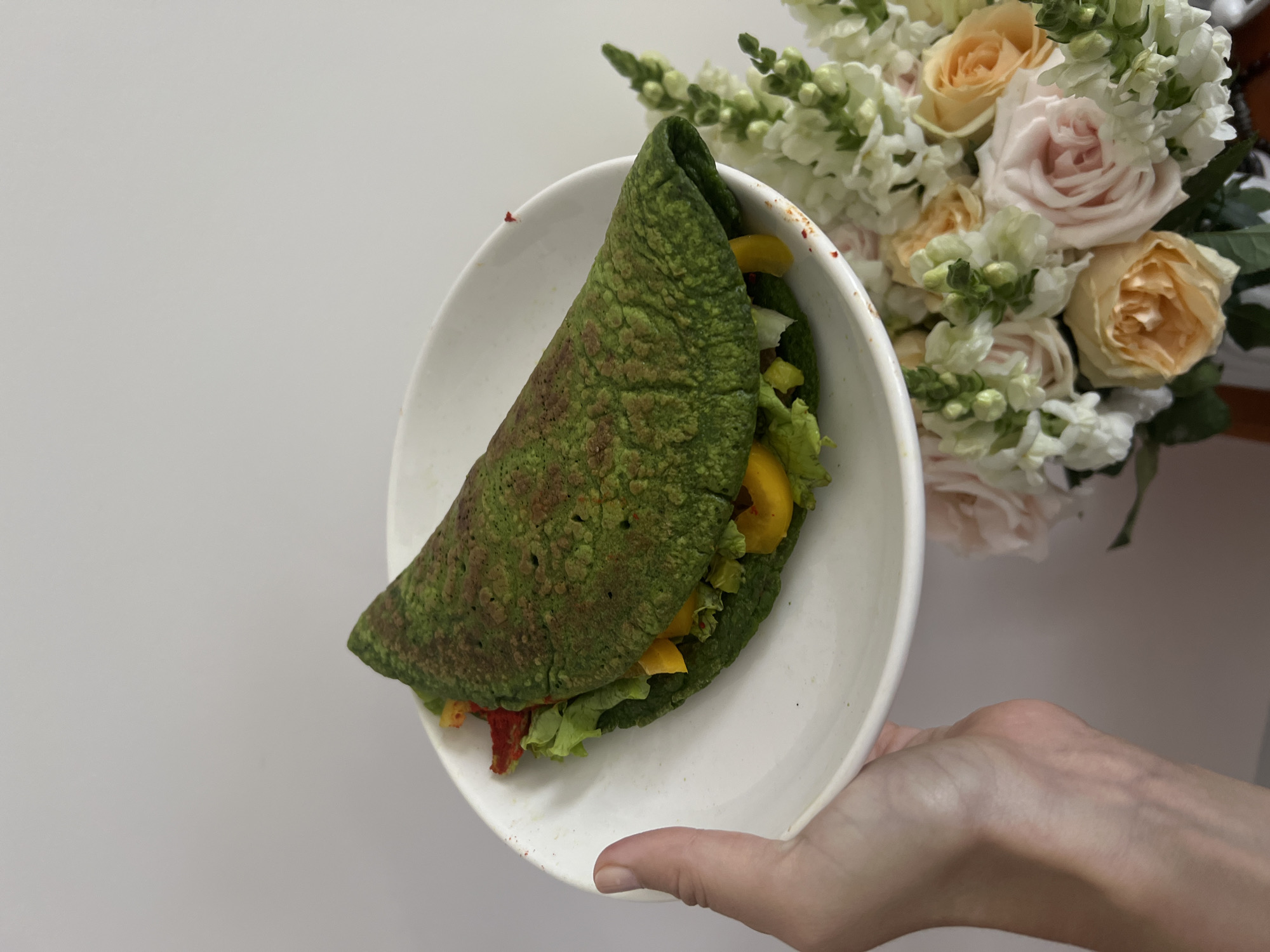 A hand holds a white bowl containing a green crepe-like wrap filled with lettuce, yellow bell peppers, and red peppers. Behind the bowl is a bouquet of light pink and peach roses with white flowers. The background is a plain, light-colored wall. The location is not discernible from the image.